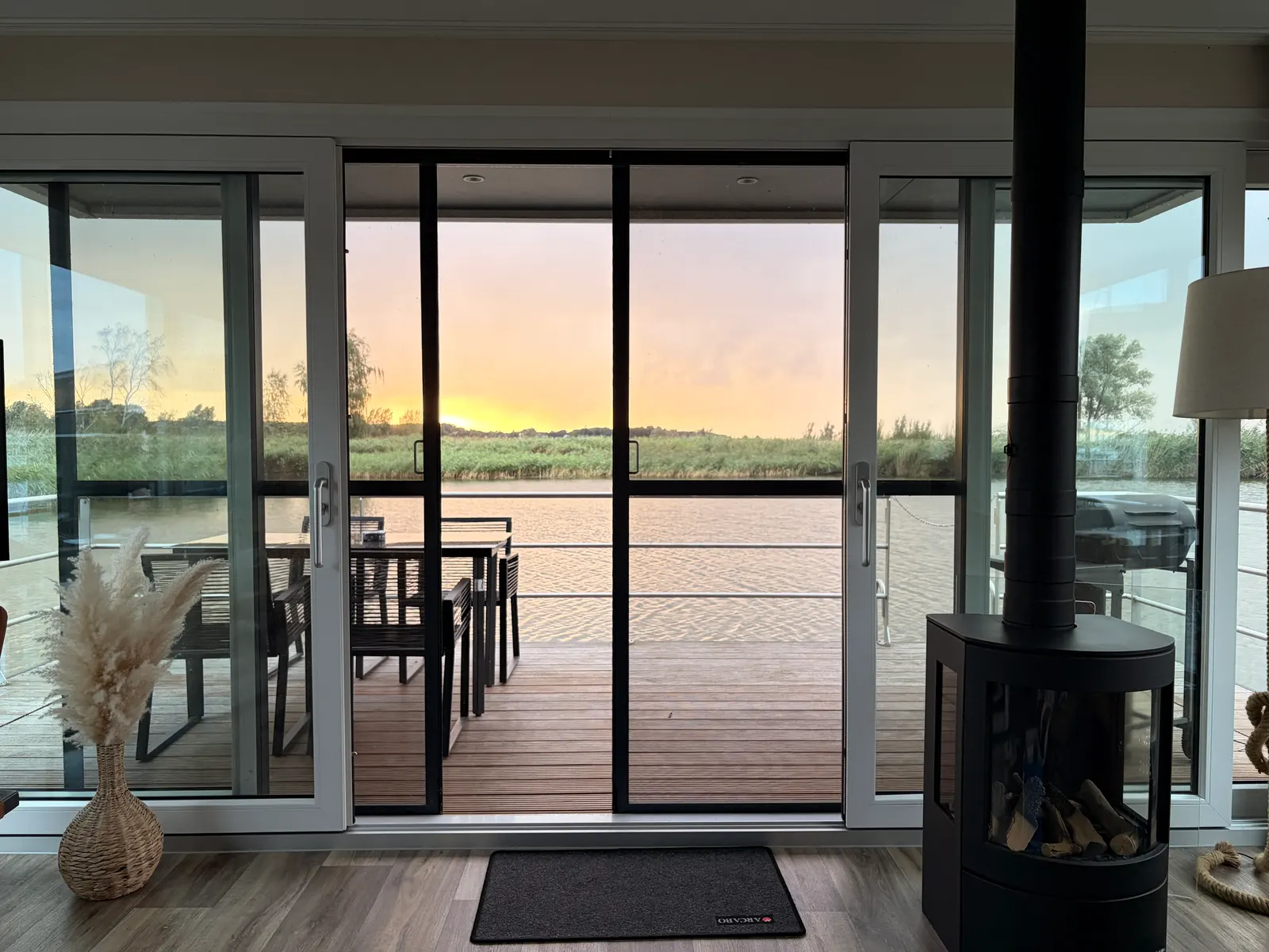 Geöffnete Glasschiebetüren führen von einem Hausboot mit Kamin auf eine Holzterrasse mit Tisch und Stühlen bei Sonnenuntergang mit Blick auf die Ostsee.