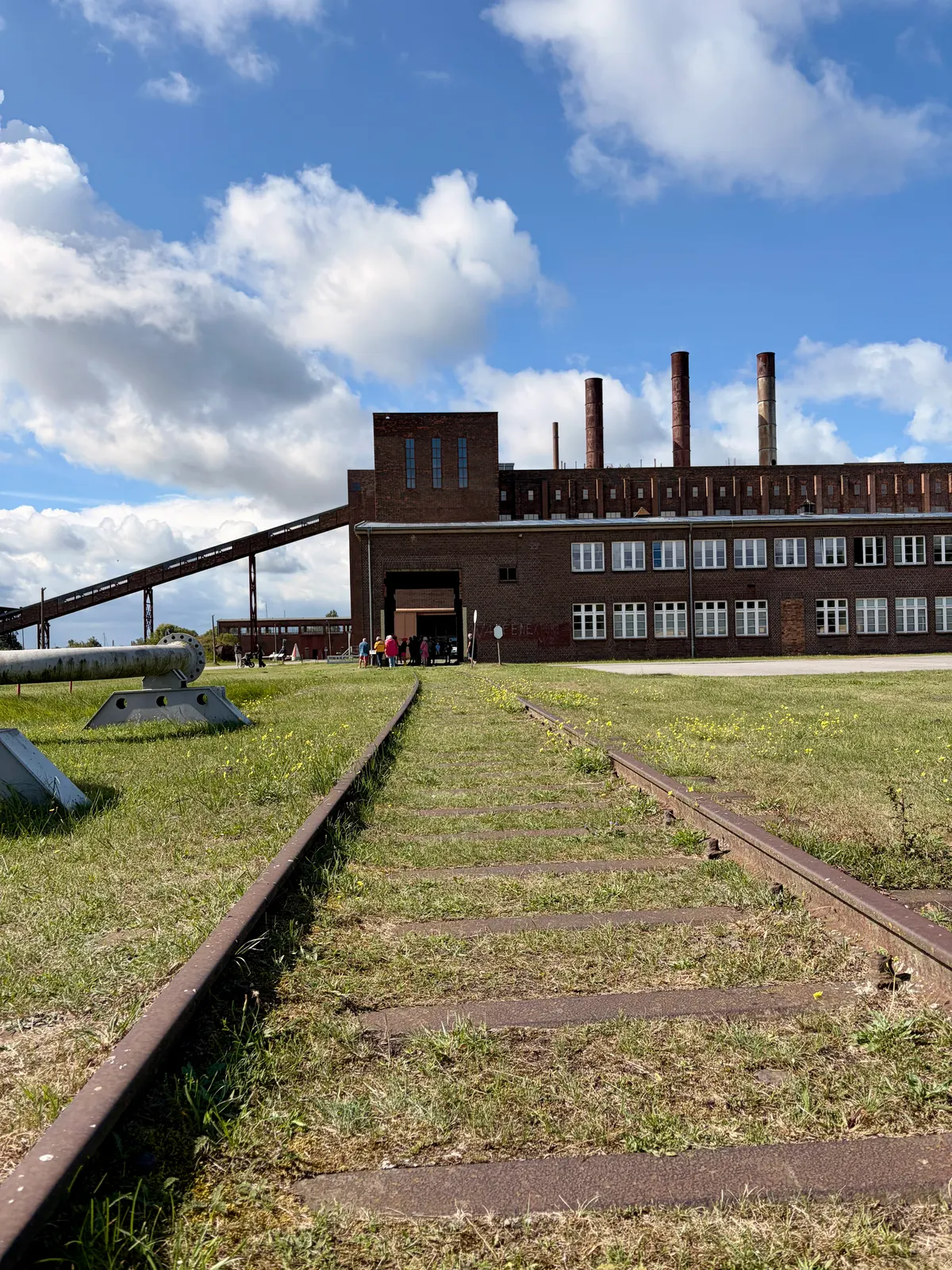 Gleise führen über eine Wiese zum ehemaligen Kohlekraftwerk des Historisch-Technischen Museums Peenemünde mit Schornsteinen und Förderband.