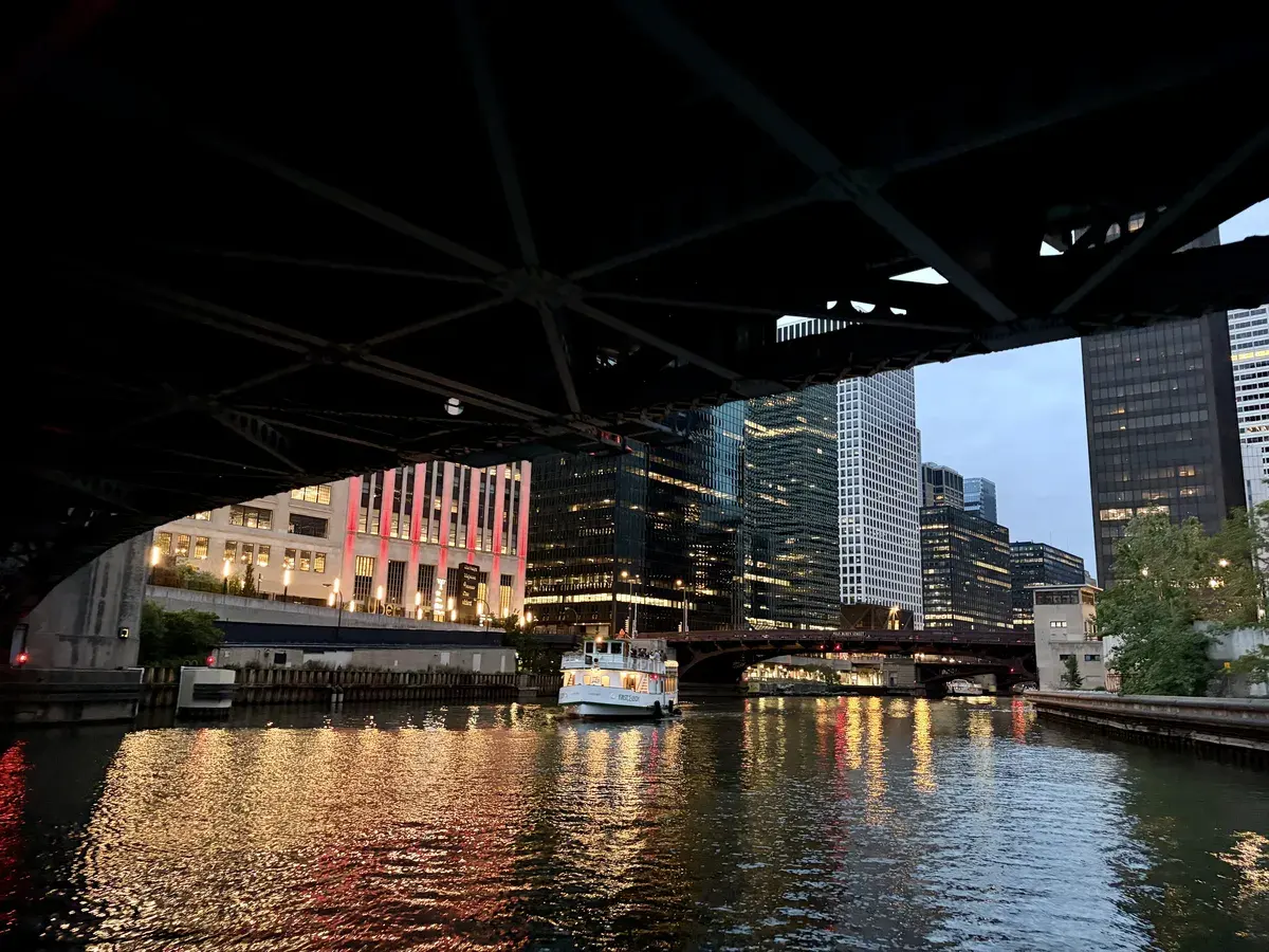 Ein Boot fährt unter einer Brücke hindurch auf einem ruhigen Fluss, der von modernen Wolkenkratzern und beleuchteten Gebäuden gesäumt ist. Die Lichter reflektieren sich im Wasser, während der Himmel in Dämmerlicht getaucht ist.