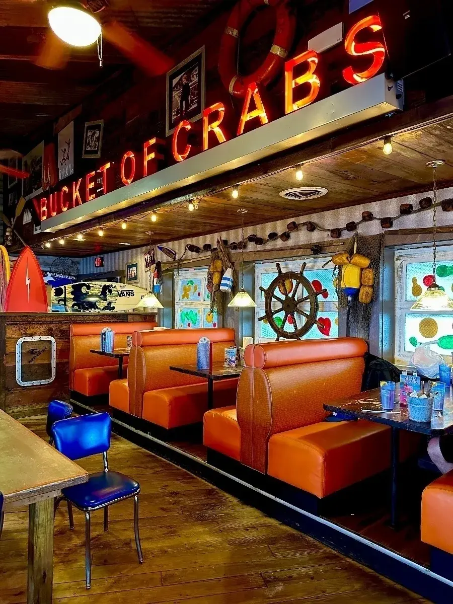 Eine Innenansicht eines rustikalen, maritim gestalteten Restaurants mit der Leuchtreklame "BUCKET OF CRABS". Es gibt orangefarbene Kunstlederbänke und Holztische, an einem Tisch steht ein blauer Stuhl.