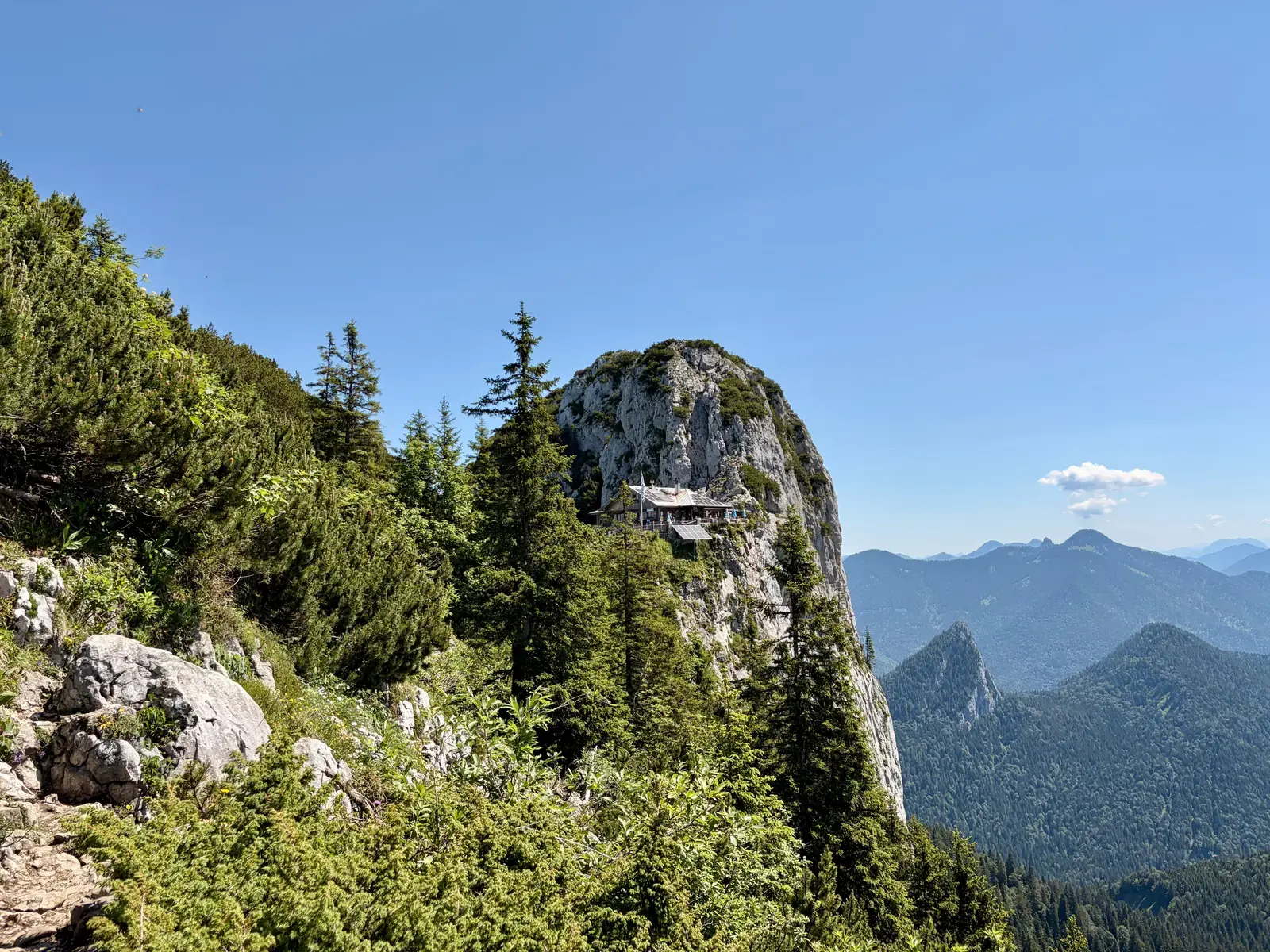 Die Tegernseer Hütte klebt wie ein Adlernest am felsigen Buchstein, umgeben von Latschen und Tannen. Dahinter reicht der Blick über die bewaldeten Gipfel der Tegernseer Berge.