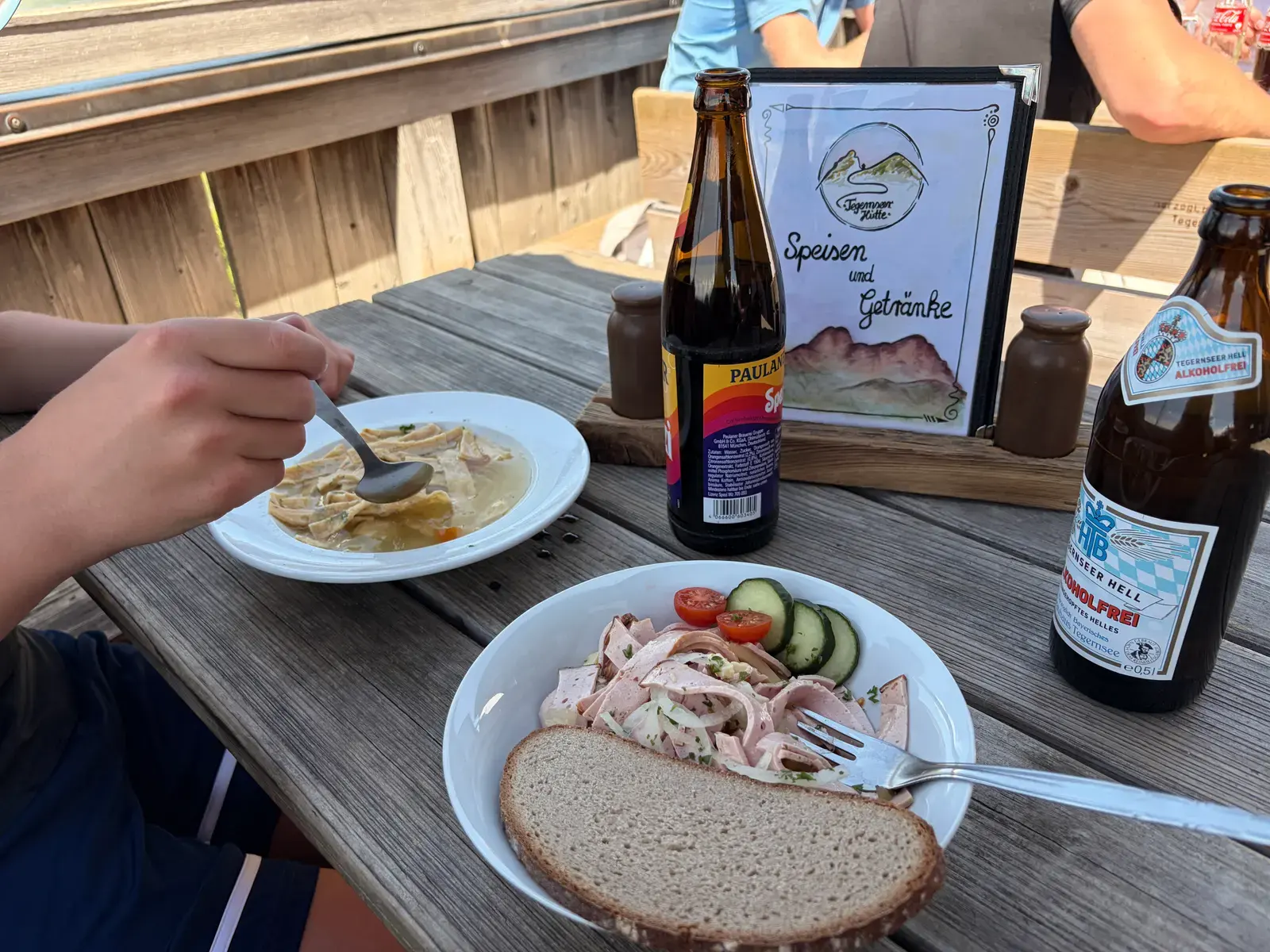 Essen und Getränke auf einem Holztisch auf der Terrasse der Tegernseer Hütte, darunter Brote, Salat und Suppe. Im Hintergrund steht die Speisekarte der Hütte.