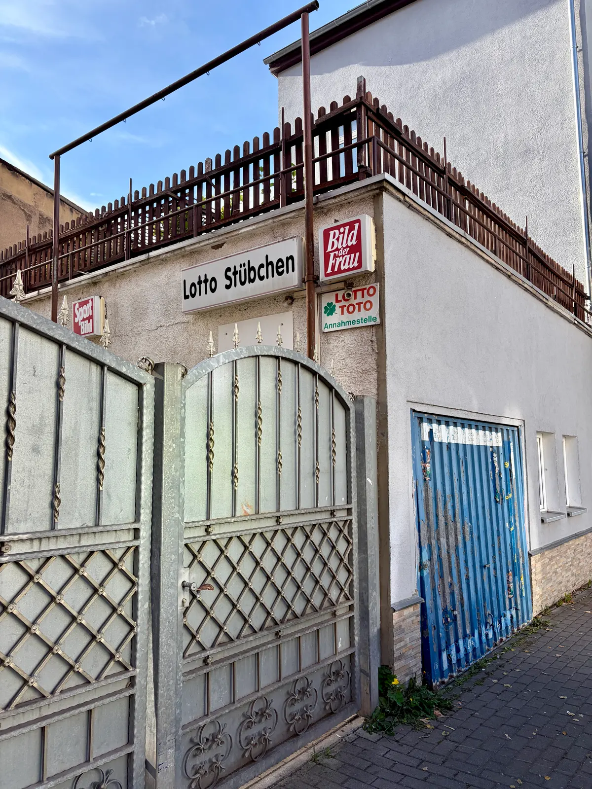 Ehemaliges Lotto-Stübchen an einer grauen Hausecke mit Metalltor, blauem Gitterfenster und Bild-der-Frau- sowie Lotto-Toto-Schildern.