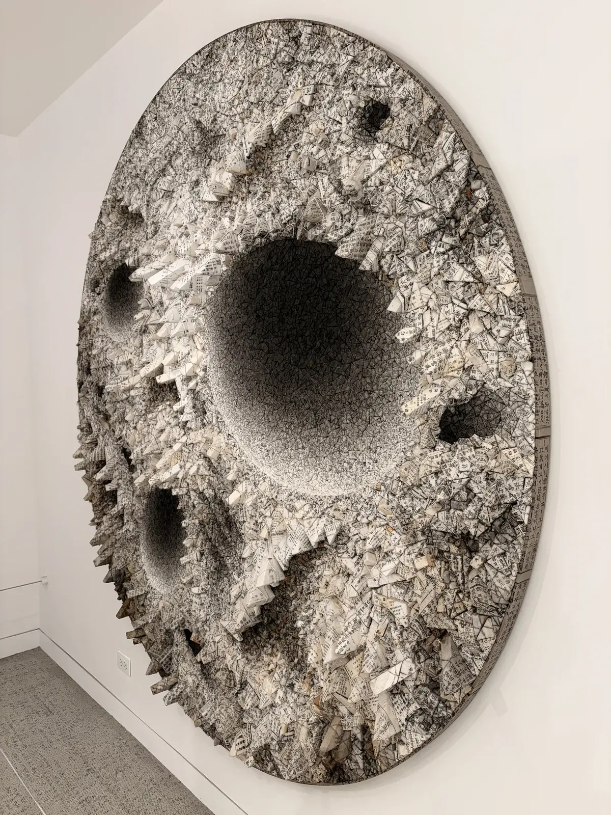 Eine große, runde Wandskulptur mit einer strukturierten Oberfläche aus grauen und weißen Materialien. Die Skulptur hat mehrere tiefe Vertiefungen und hervorstehende Formationen, die eine dreidimensionale Wirkung erzeugen.