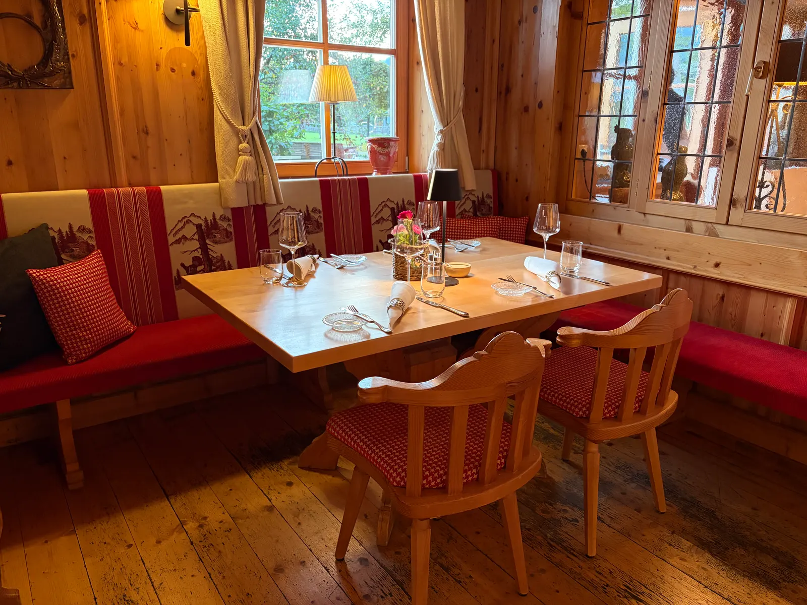 Gemütlicher Tisch in einer holzvertäfelten Hotelstube mit roten Polsterbänken, Holzstühlen, gedeckten Gläsern und Blick durch Fenster ins Grüne im Gut Steinbach