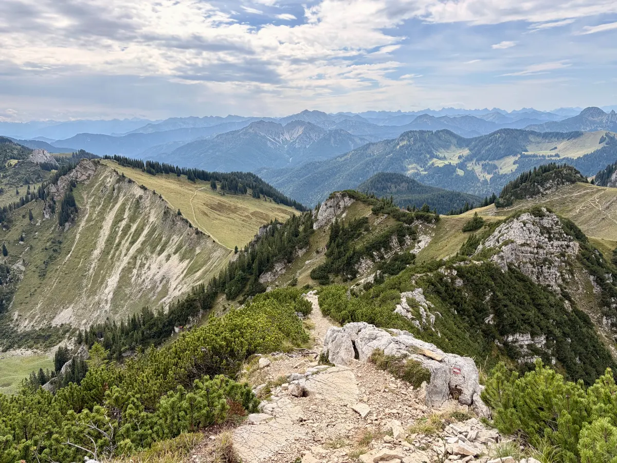 Schmaler felsiger Bergpfad auf einem Grat mit Blick auf bewaldete Gipfel und weite Alpenlandschaft unter bewölktem Himmel