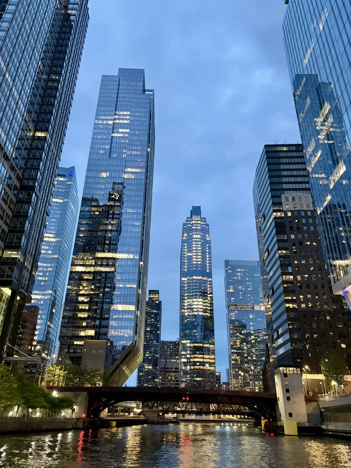Hochhäuser aus Glas spiegeln den Himmel und das Wasser eines Flusses wider, während sich auf beiden Seiten lebhafte Lichter der Stadt reflektieren. Eine Brücke spannt sich über den Fluss, der in der Dämmerung ruhig fließt.