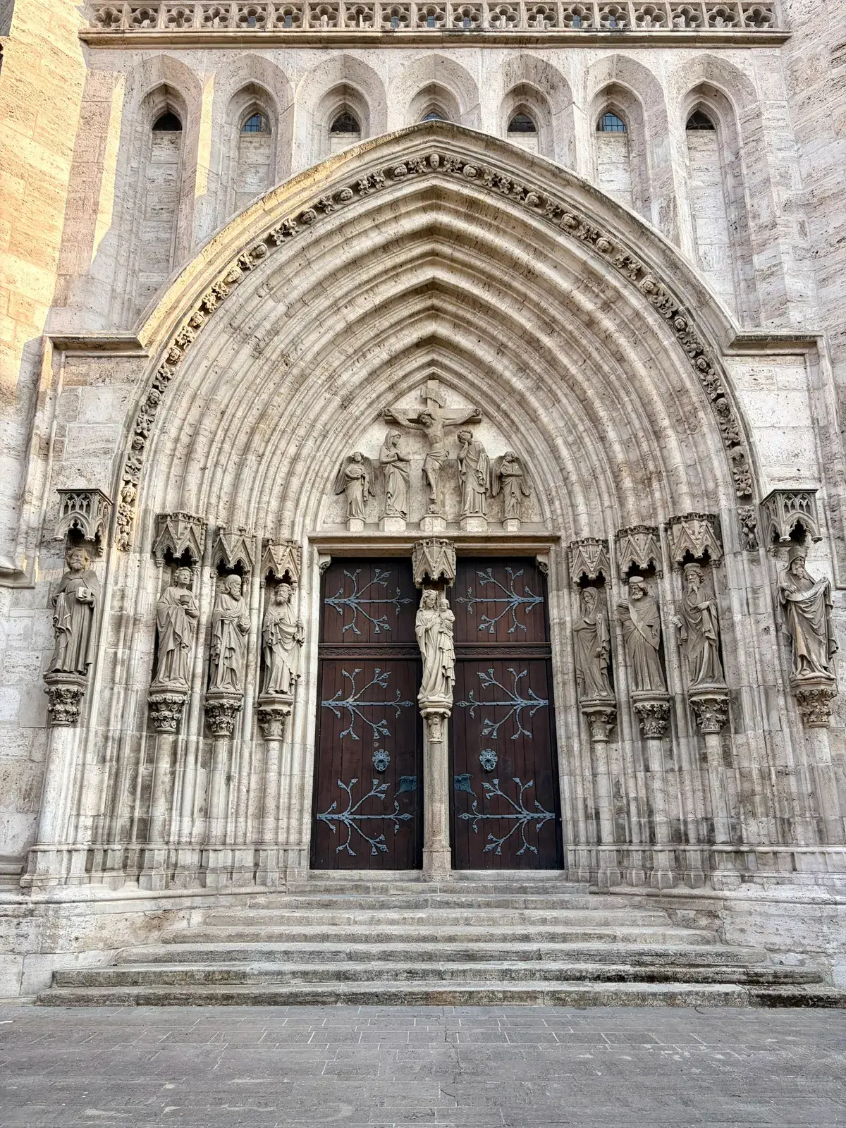 Gotisches Kirchenportal mit reich verzierten Steinbögen, Figuren und einer dunklen zweiflügeligen Tür. Davor führen mehrere breite Stufen zum Eingang hinauf.