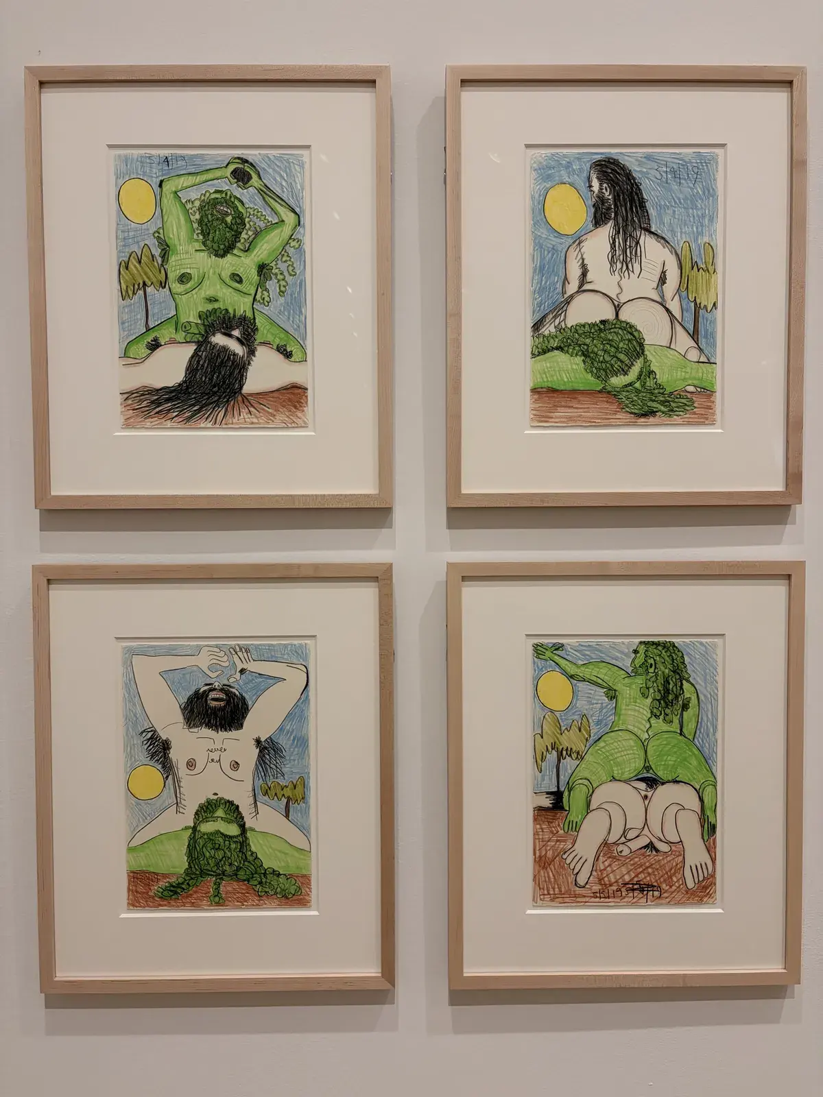 Vier gerahmte Illustrationen sind an einer Wand aufgehängt, die verschiedene Figuren in einem natürlichen Umfeld darstellen. Die Charaktere sind mit grünen und braunen Farben gestaltet und zeigen intime Posen zwischen Bäumen und einer gelben Sonne.