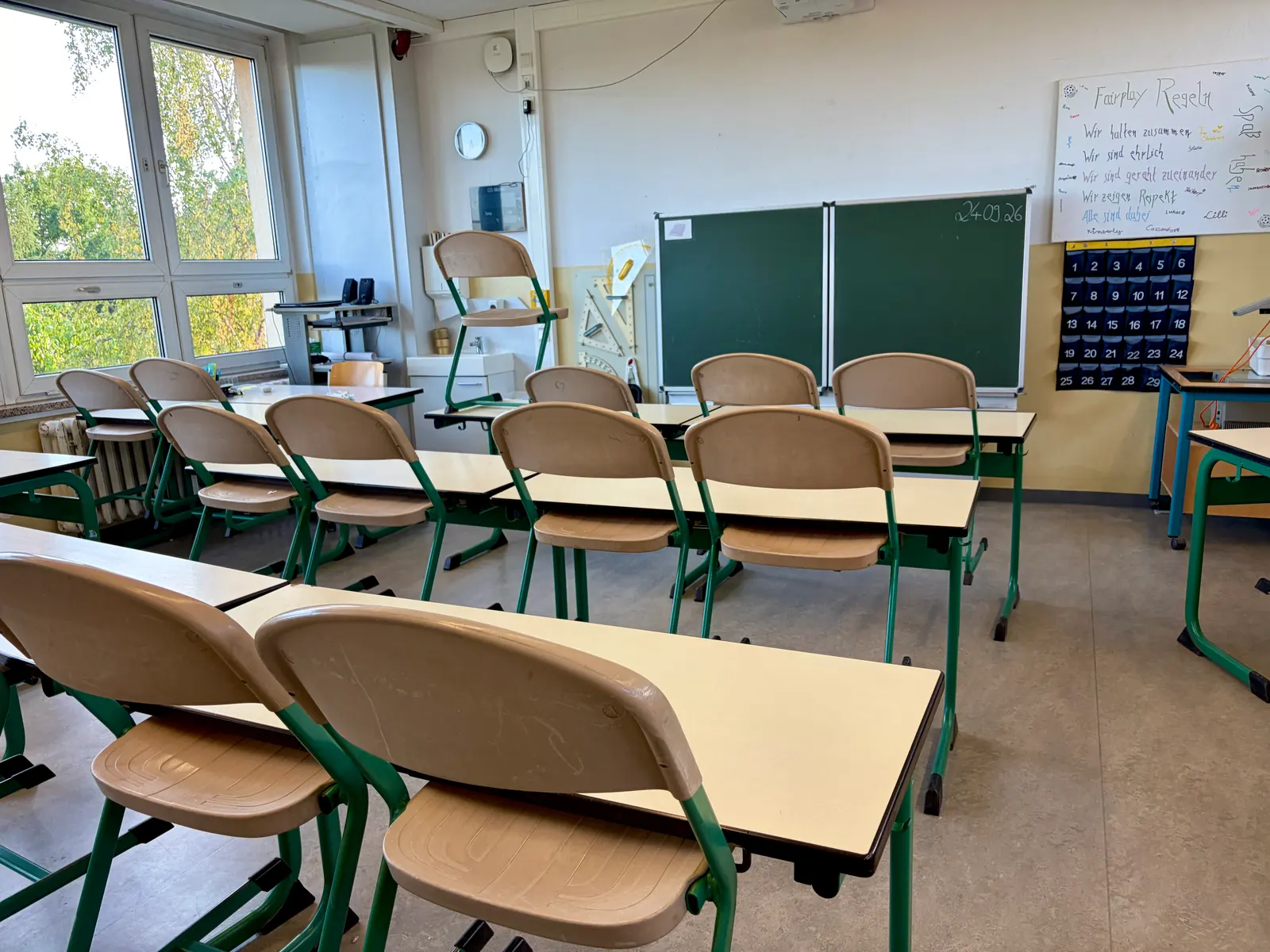 Klassenzimmer mit Reihen aus grünen Tischen und braunen Stühlen, grüner Tafel, Flipchart und großen Fenstern mit Blick ins Grüne