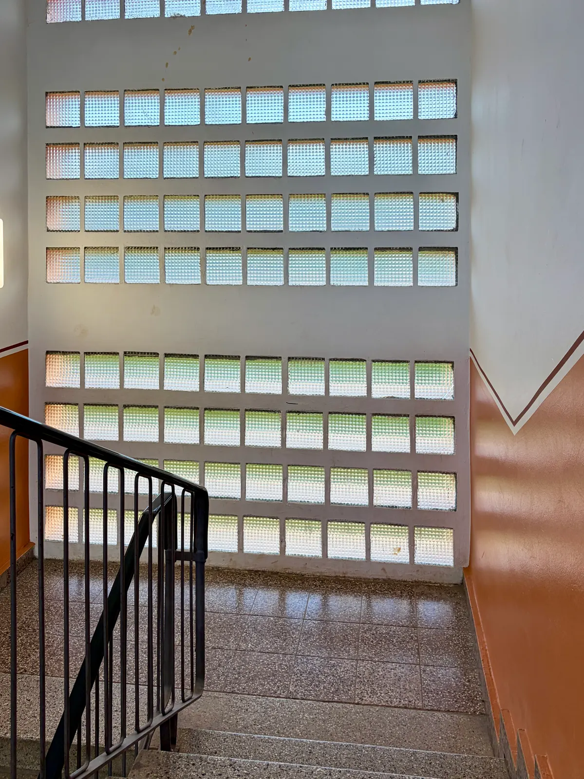 Treppenhaus mit Glasbausteinwand, braun gestrichenen Wänden, schwarzem Geländer und Steinfußboden