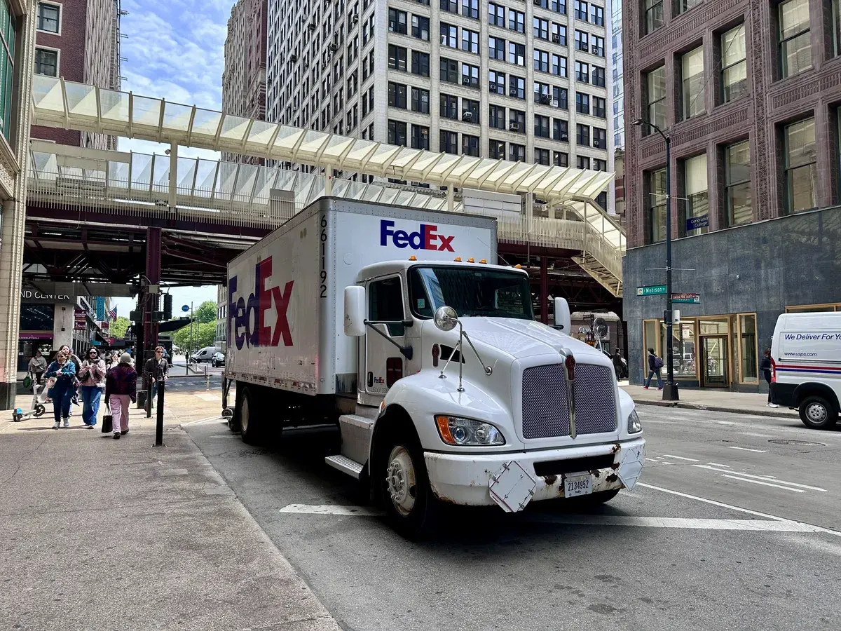 Ein weißer FedEx-Lkw steht auf einer städtischen Straße, umgeben von hohen Gebäuden. Fußgänger gehen auf dem Bürgersteig vorbei, während eine Überführung in der Nähe sichtbar ist.