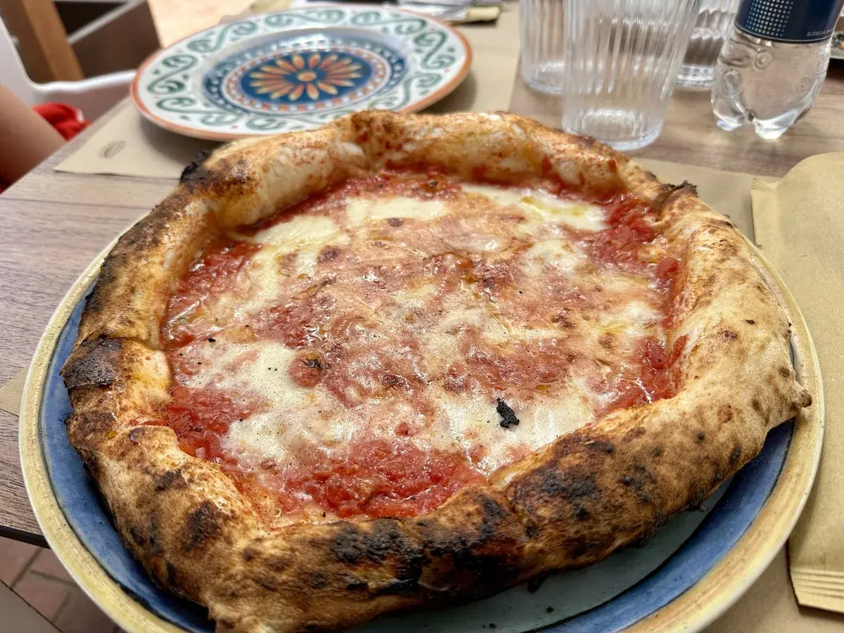 Eine Pizza mit Tomatensoße und geschmolzenem Käse liegt auf einem Teller auf einem Tisch, umgeben von Gläsern und einem anderen Teller mit Muster.