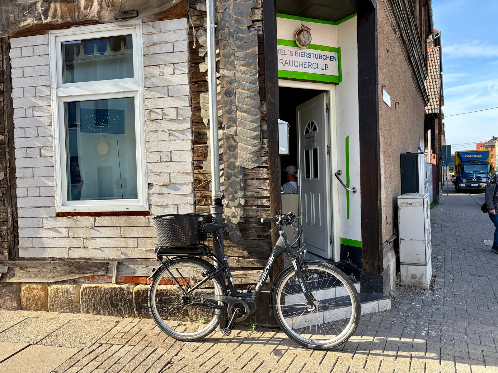 Fahrrad vor einem Hauseingang mit bröckelnder Fassade und Schaufenster; im Hintergrund Straße und Müllfahrzeug.