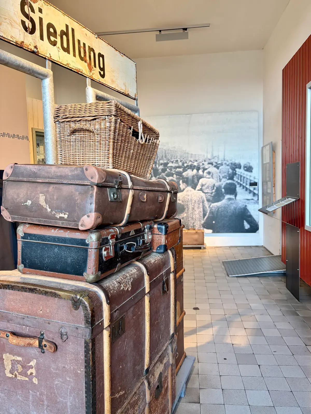 Gestapelte alte Koffer und ein Weidenkorb in einer Museumsausstellung zur „Siedlung“, im Hintergrund eine historische Schwarzweißaufnahme von Menschen am Wasser