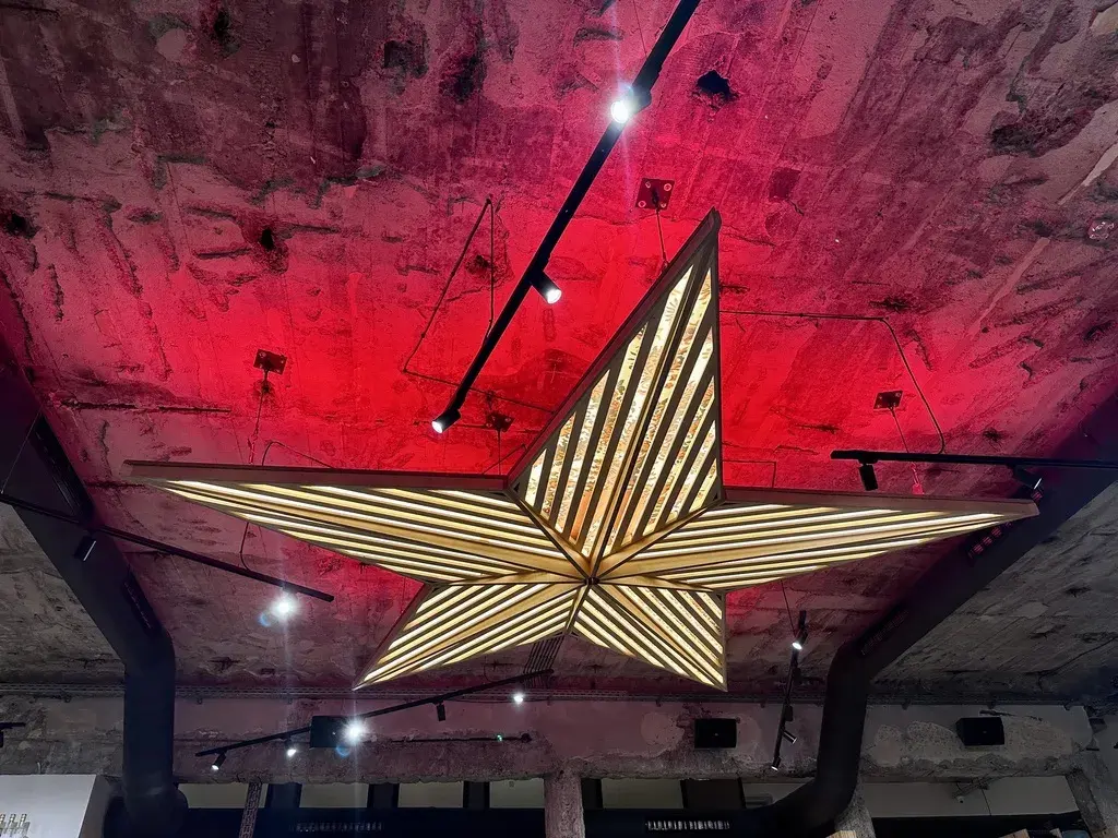 Eine große, hängende Sternleuchte mit geometrischem Design aus Holz und vielen kleinen Lichtern strahlt in einem Raum mit rotem Licht und sichtbaren Betonwänden.