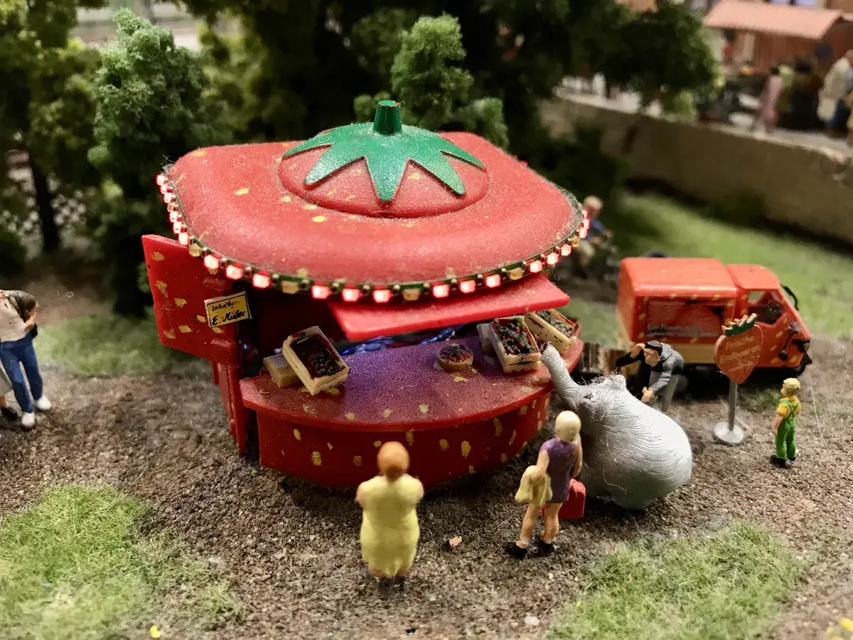 Nahaufnahme eines Miniatur-Erdbeerkiosks auf einer Modellbauanlage. Der rote, erdbeerförmige Verkaufsstand ist beleuchtet und wird von kleinen Figuren sowie einem Miniatur-Ottifanten besucht.