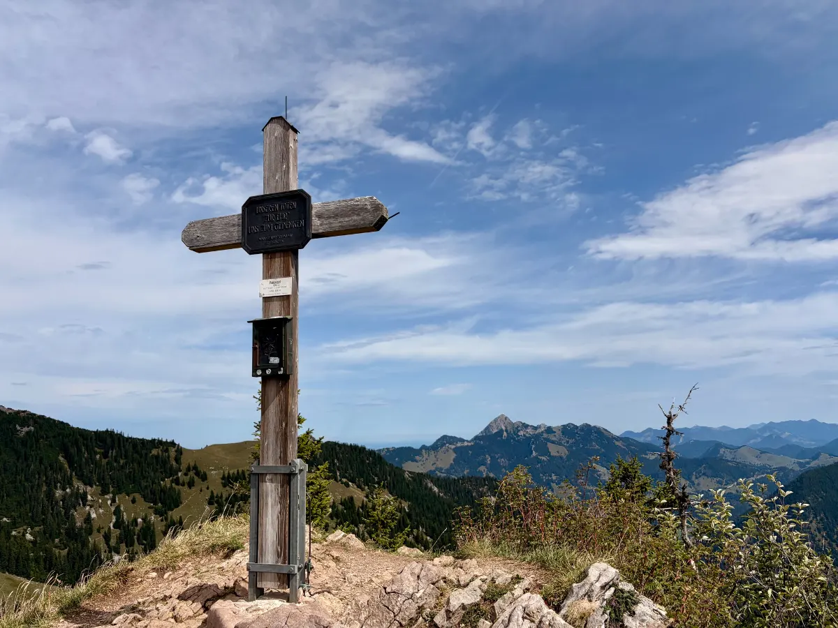 Gipfelkreuz auf felsigem Berggrat mit Blick auf bewaldete Hänge und die Alpen unter blauem, leicht bewölktem Himmel