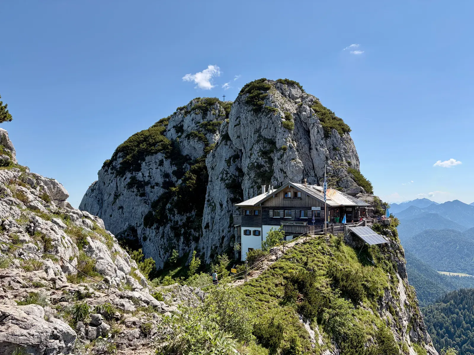 Tegernseer Hütte auf einem schmalen Felsgrat unter dem Buchstein, umgeben von steilen Kalkfelsen und weitem Bergpanorama bei klarem Himmel.