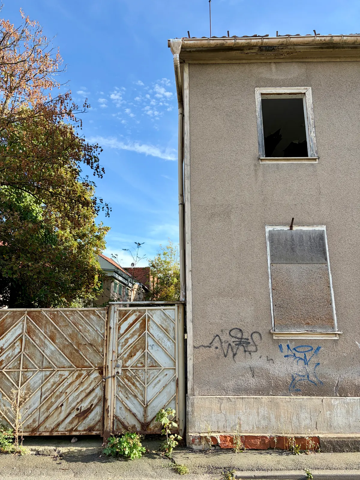 Vernageltes Hoftor neben einer grauen Hausfassade mit beschädigten Fenstern, Graffiti und überwuchertem Randstreifen unter blauem Himmel