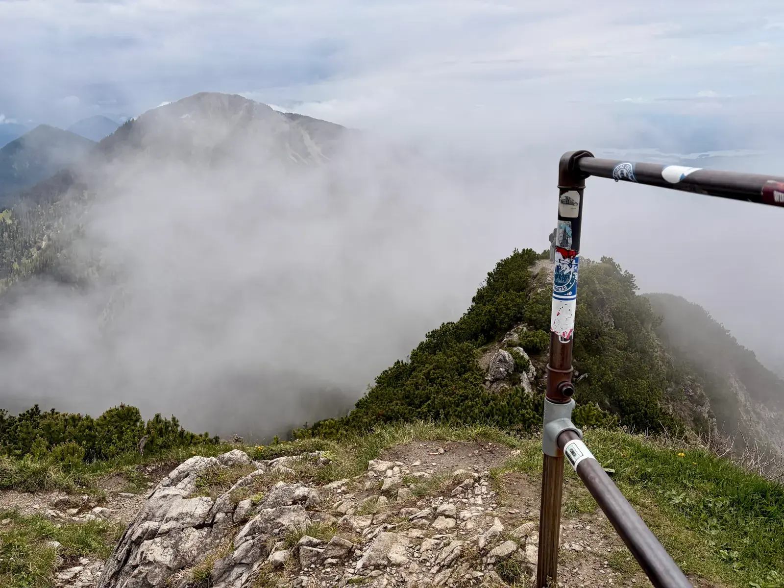 Nebel zieht über den felsigen Grat zwischen Heimgarten und Herzogstand, ein Geländer markiert den schmalen Weg. Die umliegenden Berggipfel sind nur schemenhaft durch die Wolken zu sehen.