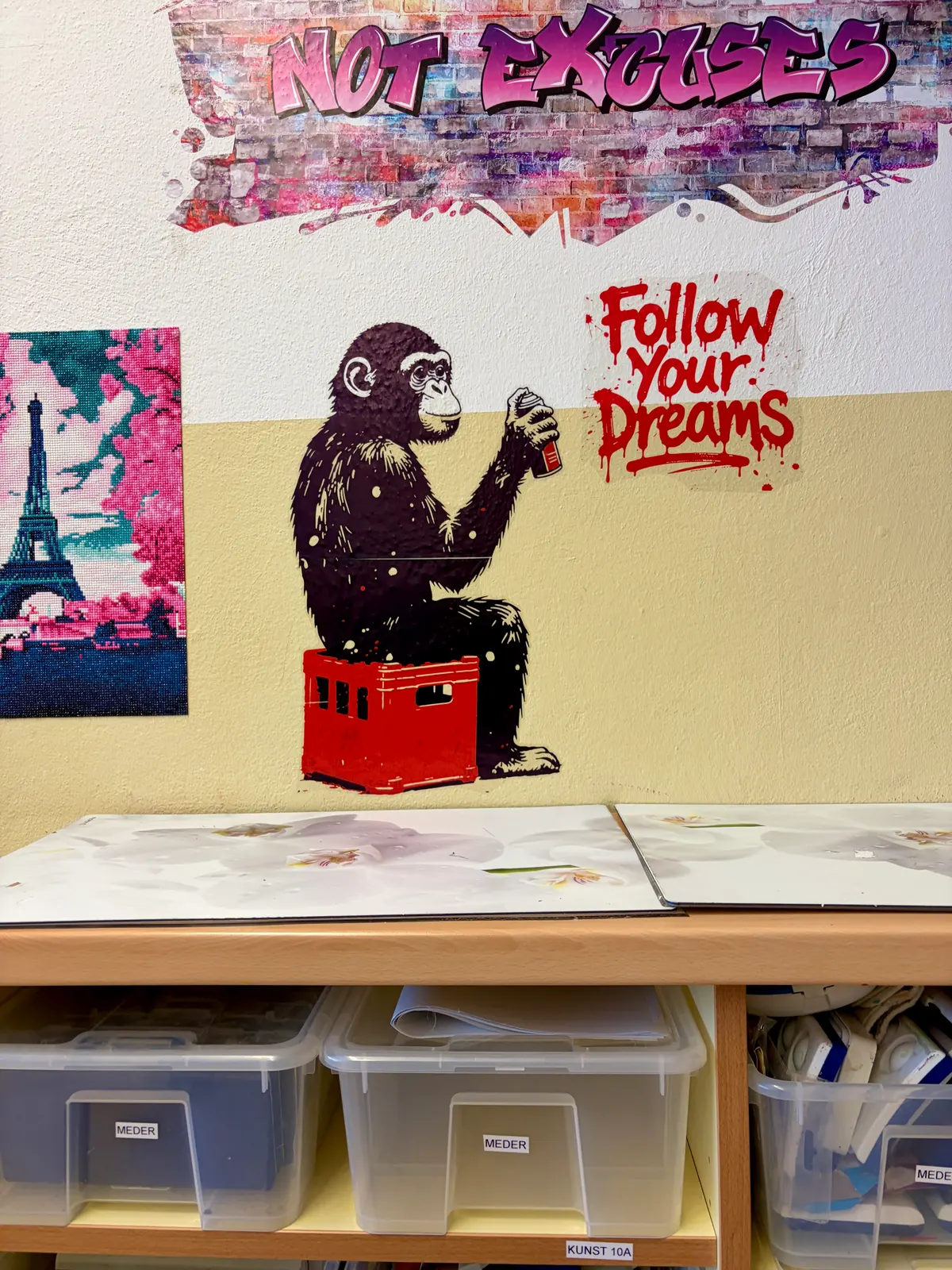Wand in einem Klassenraum mit Street-Art-Bildern und Sprüchen, darunter Regale mit beschrifteten Materialboxen und ein Tisch mit aufgeschlagenen Mappen