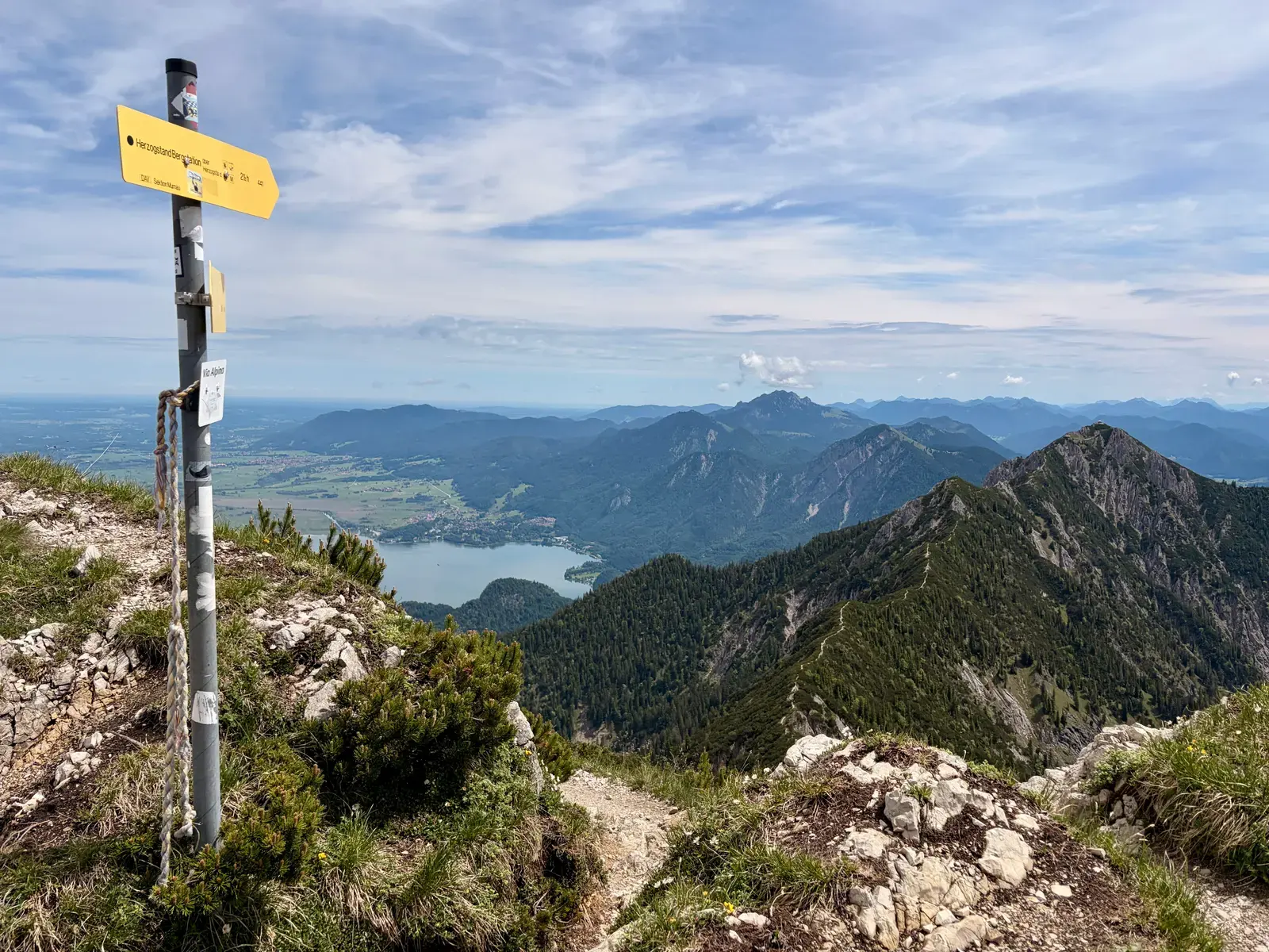 Gelber Wegweiser auf einem felsigen Gipfelgrat auf dem Heimgarten zeigt in Richtung Herzogstand. Dahinter reicht der Blick über Kochlsee Walchensee und die umliegenden Berge.