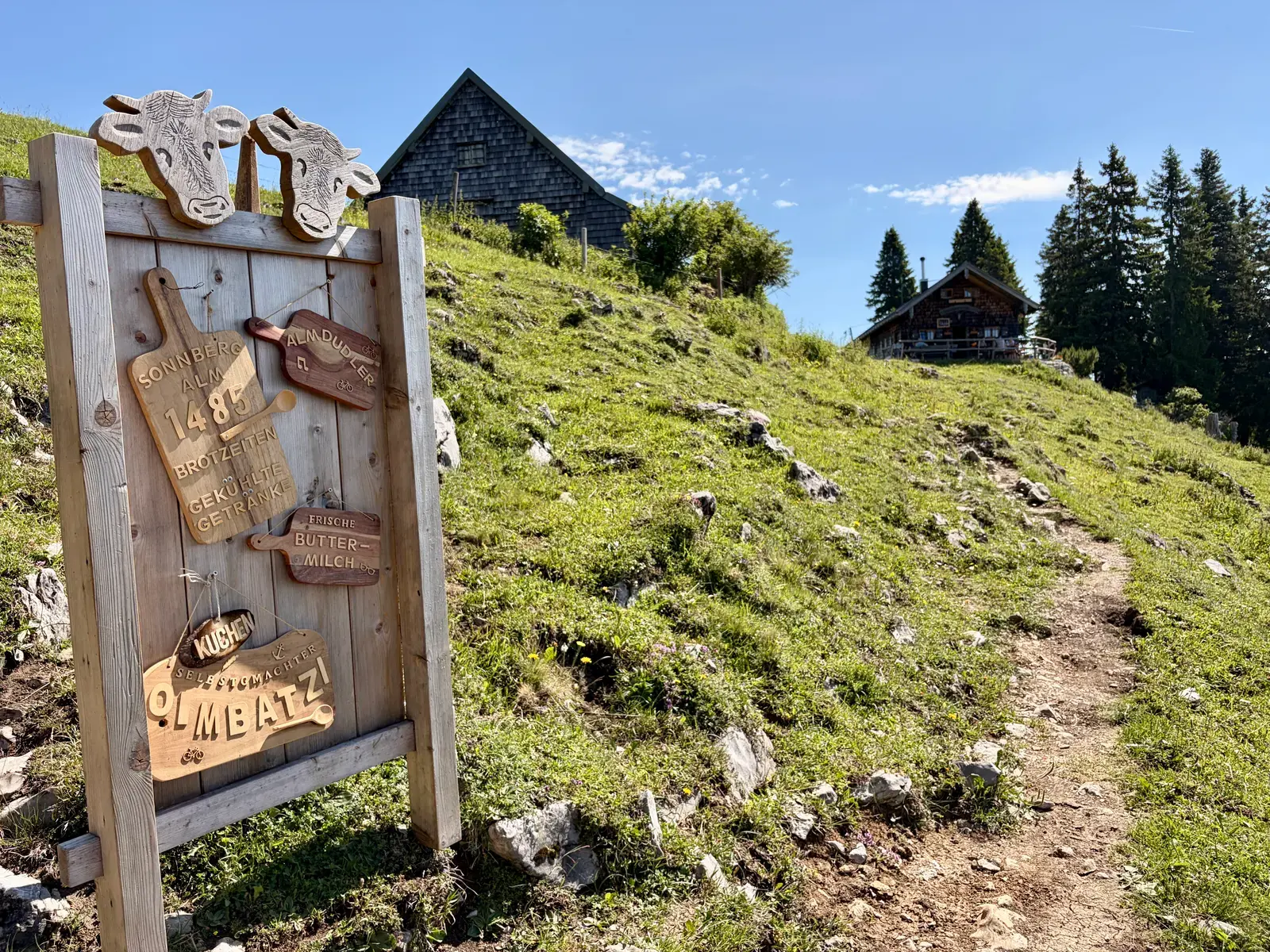 Holzschild der Sonnbergalm am Bergweg zur Tegernseer Hütte, daneben Almwiesen und Hütten unter blauem Himmel.