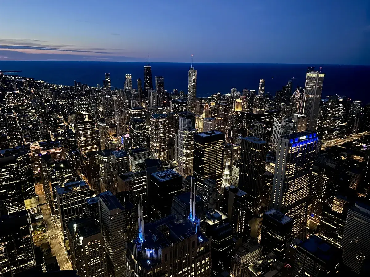 Eine beeindruckende Skyline von Chicago bei Dämmerung, mit zahlreichen beleuchteten Wolkenkratzern und der ruhigen Seeoberfläche im Hintergrund. Die Lichtreflexe der Gebäude sorgen für eine lebendige Atmosphäre.