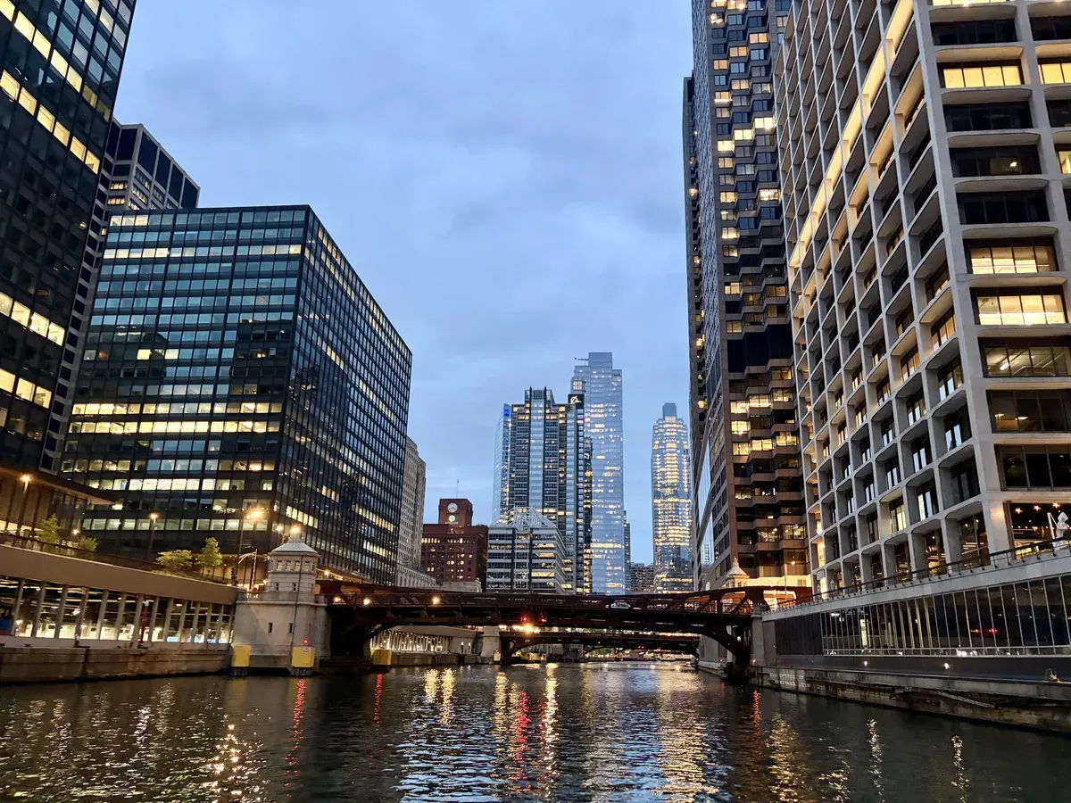 Eine Stadtansicht bei Dämmerung, die einen Fluss zeigt, umgeben von modernen Wolkenkratzern. Die reflektierenden Lichter der Gebäude sind im Wasser sichtbar, während eine Brücke den Fluss überspannt.