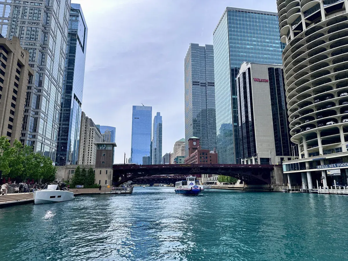 Die Szene zeigt einen Blick auf einen Stadtfluss, der von modernen Wolkenkratzern flankiert wird. Eine Brücke überspannt den Fluss, während ein Boot vorbeifährt und Bäume entlang der Uferpromenade sichtbar sind.