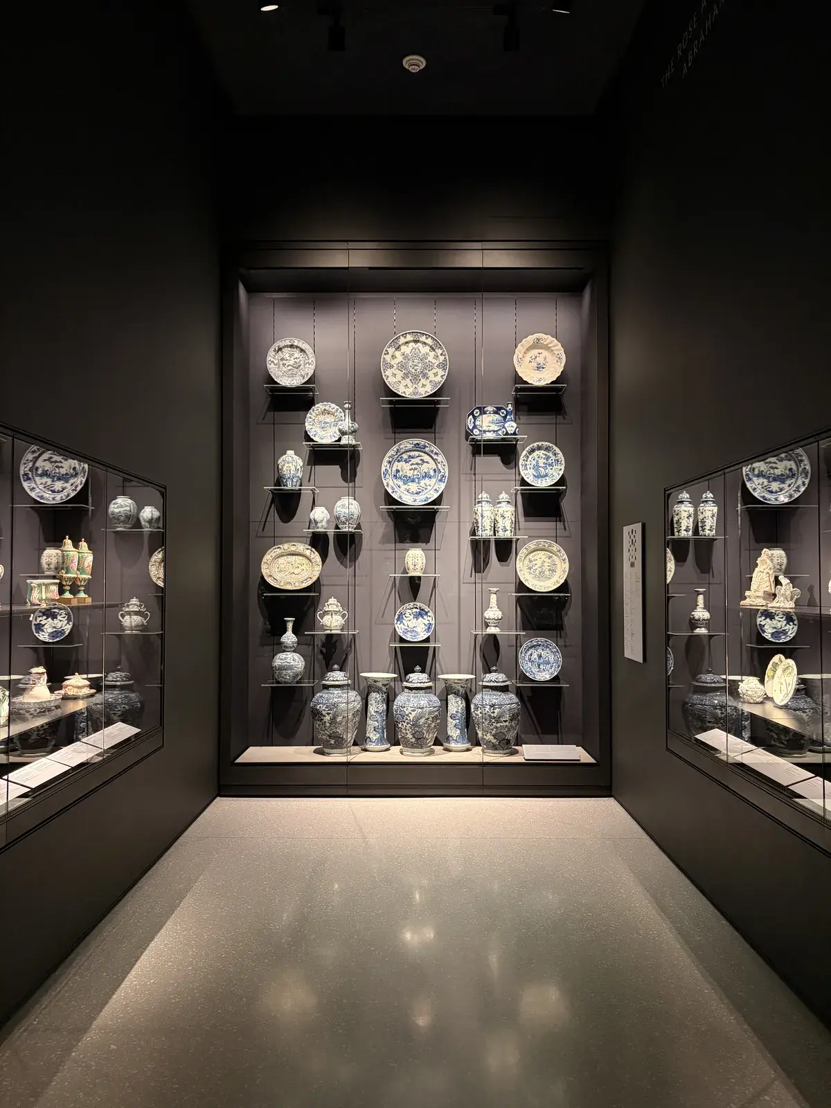 Eine dunkle Museumshalle mit zwei Vitrinen, die zahlreiche historische Porzellanobjekte zeigen. An der Wand hängen große Teller und Vasen mit blau-weißen Mustern, während die Vitrinen kleinere ornamente und Töpferwaren präsentieren.