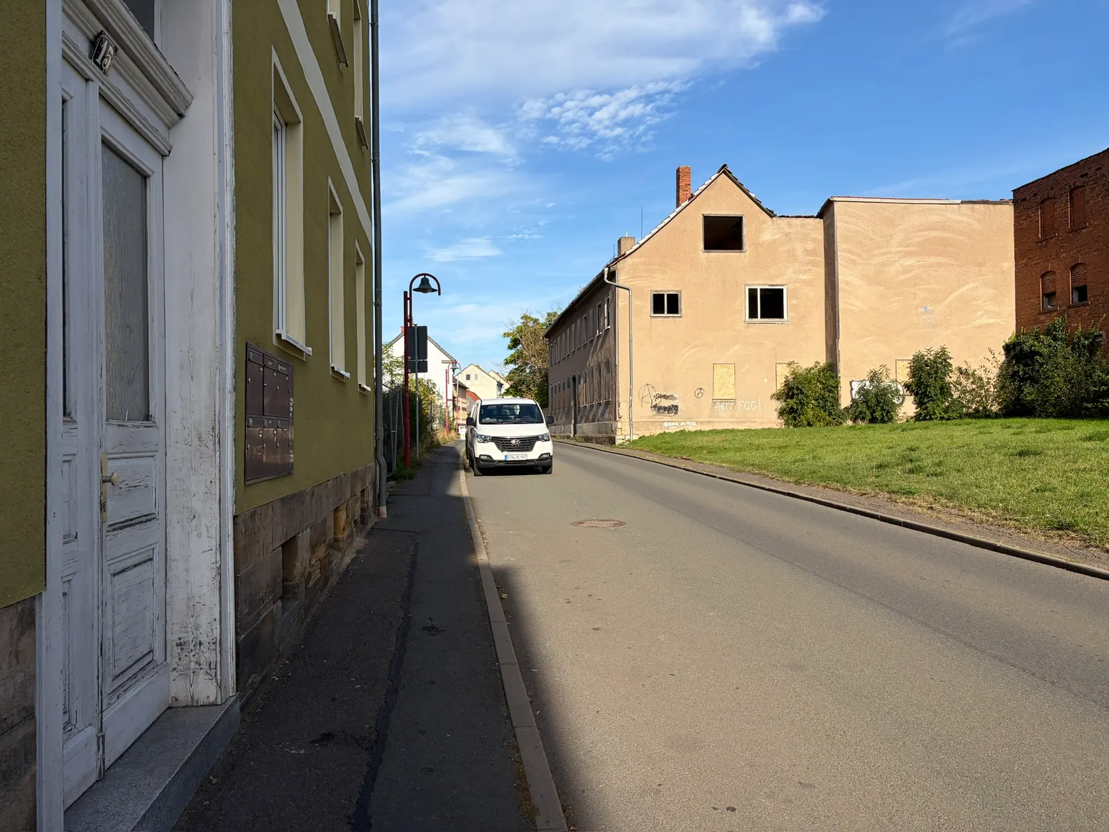 Schmale Straße in Gotha mit alten Wohnhäusern, Gehweg, Grünfläche und einem weißen Transporter unter blauem Himmel