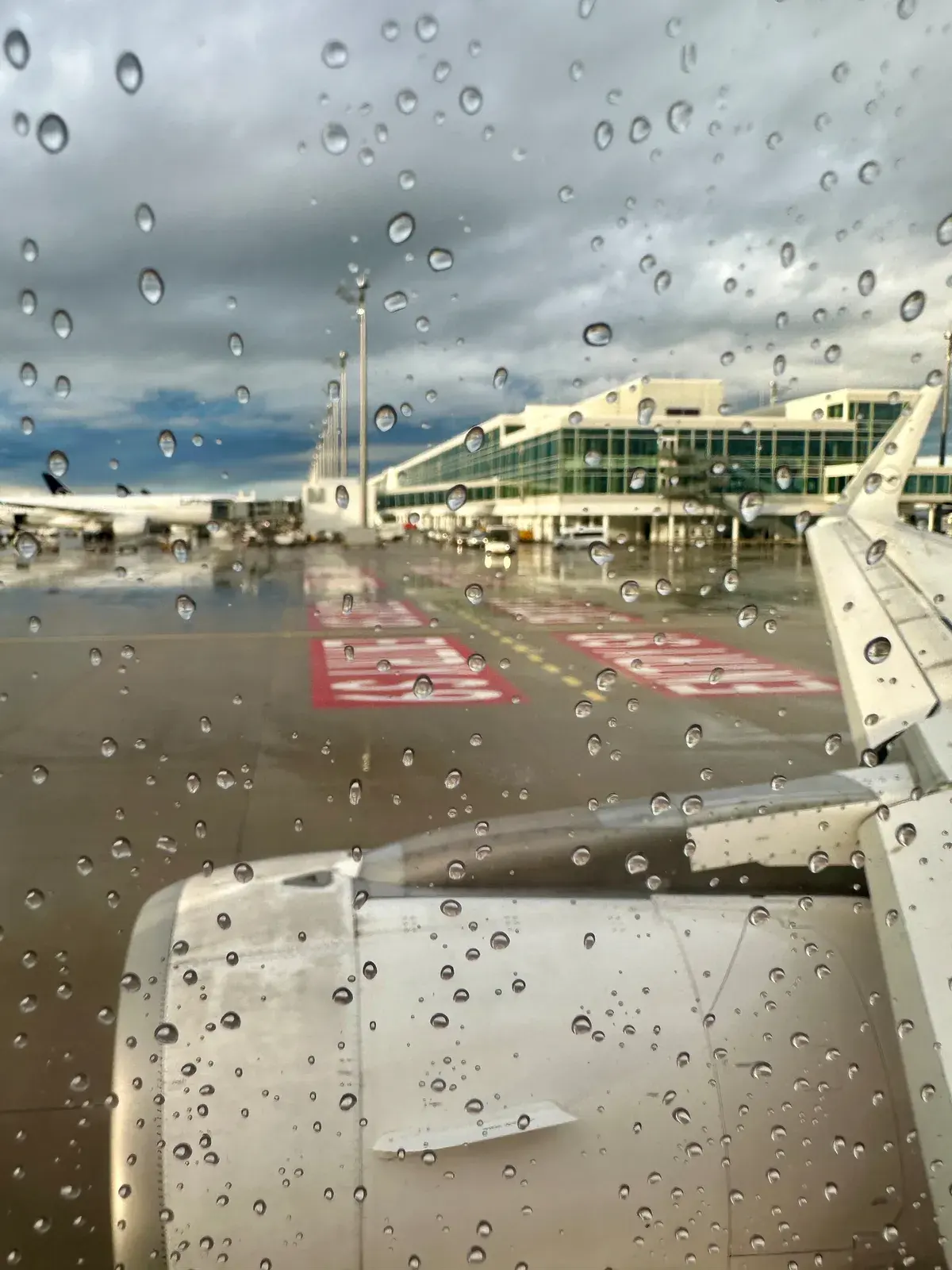 Ein Flugzeugflügel und ein Triebwerk sind durch ein regennasses Fenster auf einem Flughafen mit Terminalgebäuden und bewölktem Himmel sichtbar.