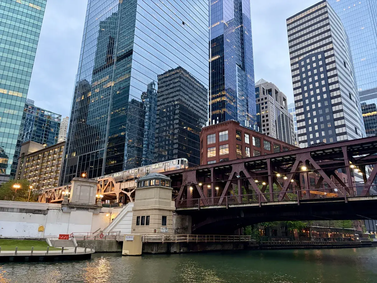 Eine U-Bahn fährt über eine Brücke aus rotbraunem Stahl, die über einen schmalen Kanal verläuft. Umgeben von modernen Wolkenkratzern aus Glas und Stahl, spiegelt sich die Architektur im Wasser wider.
