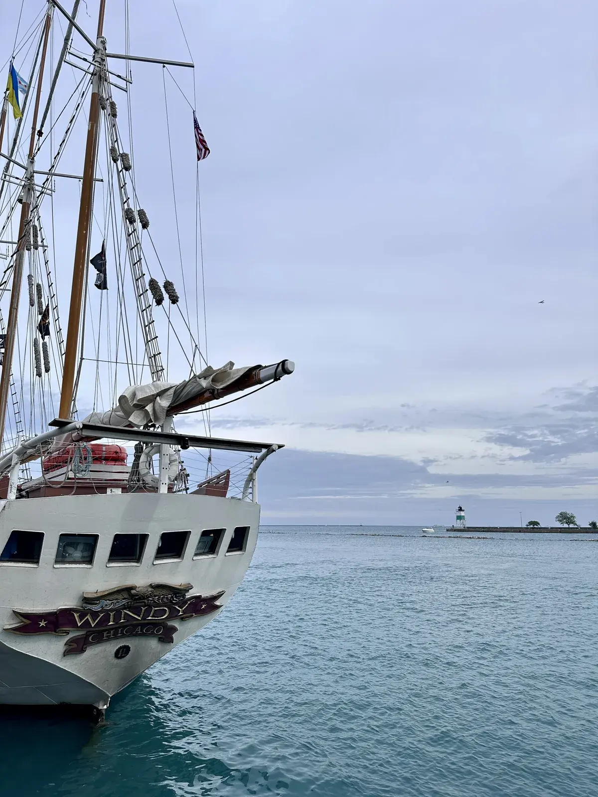 Eine Segelschiff mit mehreren Masten liegt nahe am Ufer eines ruhigen Gewässers. Im Hintergrund ist ein kleiner Leuchtturm und bewölkter Himmel zu sehen.