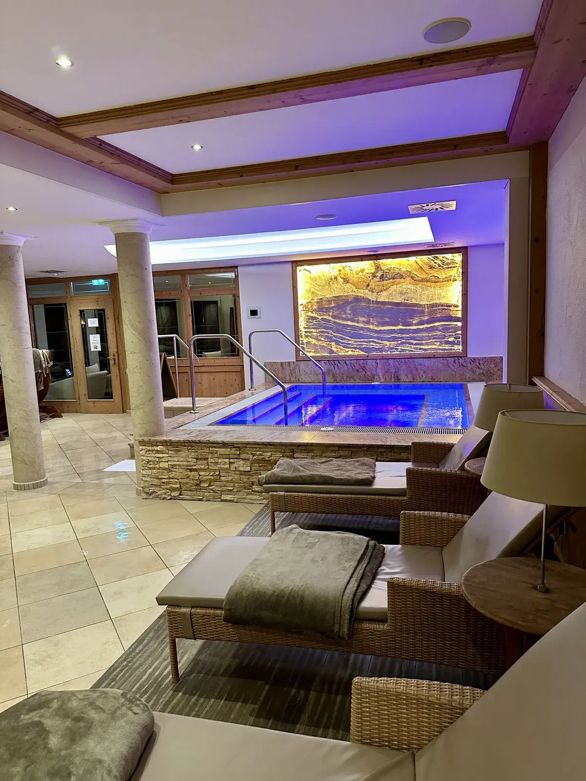 Ein moderner Wellnessbereich mit einem kleinen Pool, umgeben von Steinwänden und beleuchtet mit sanftem, violettem Licht. Im Vordergrund sind Liegen mit grauen Decken und ein Tisch mit einer Lampe zu sehen.