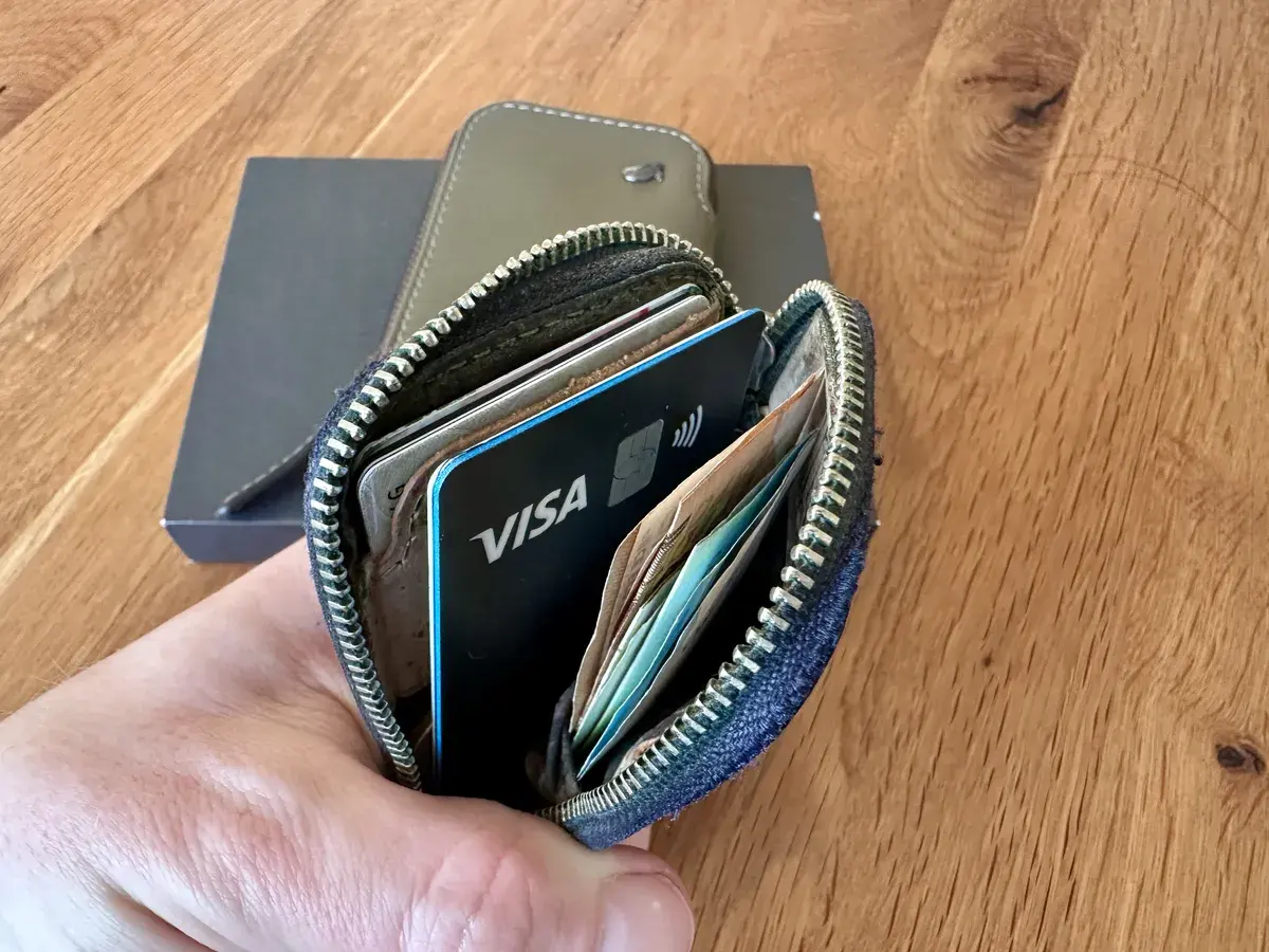 Eine Hand hält ein offenes Portemonnaie, das eine Visa-Karte und mehrere Geldscheine sowie weitere Karten sichtbar zeigt. Im Hintergrund ist eine dunkle Box aus Karton zu sehen, auf einem Holztisch platziert.