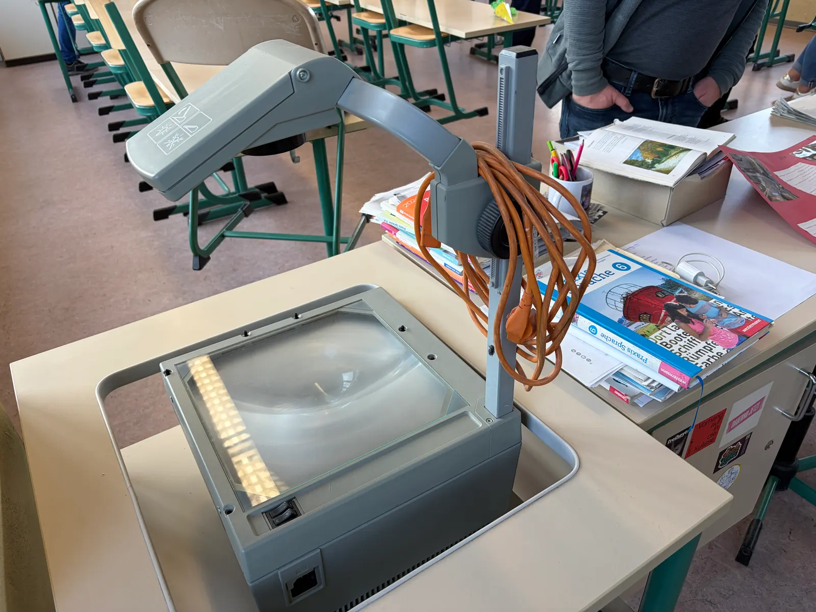 Alter Overheadprojektor auf einem Schultisch, daneben Hefte und Stifte in einem Klassenraum.
