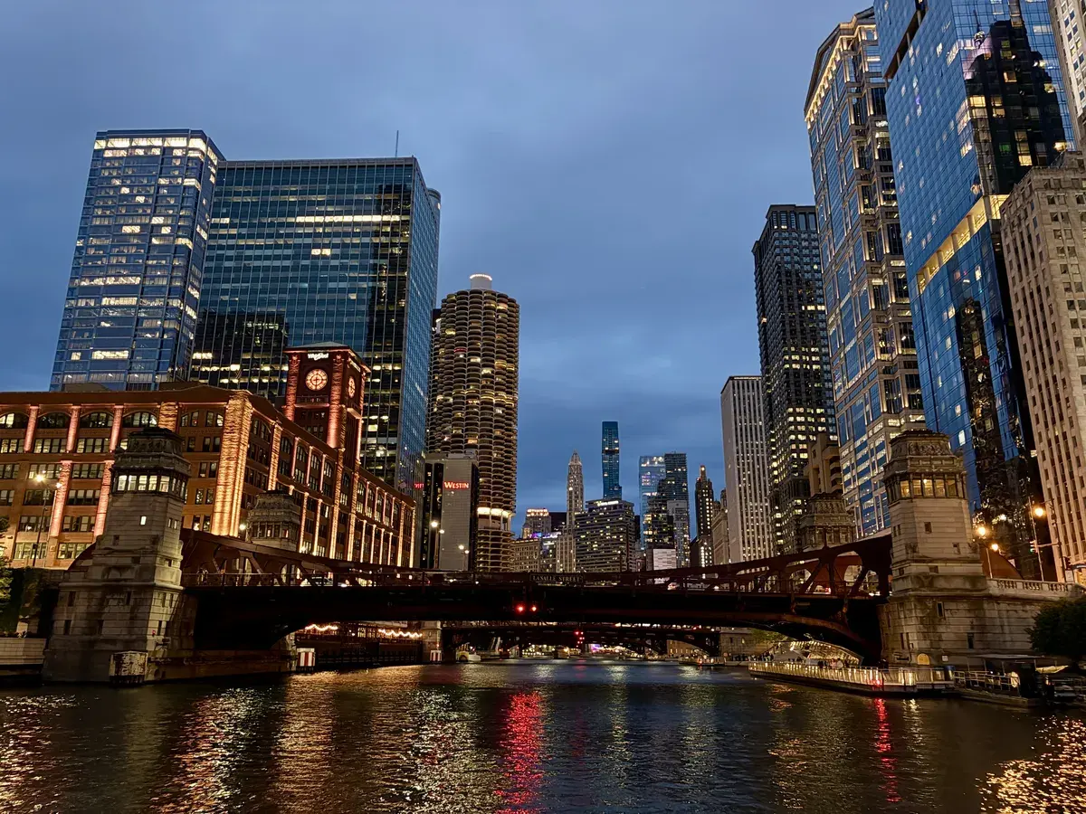 Die Skyline von Chicago bei Dämmerung, mit hohen, gläsernen Wolkenkratzern und einem beleuchteten Brückenbogen über dem ruhigen Fluss, dessen Wasser die Lichter reflektiert.
