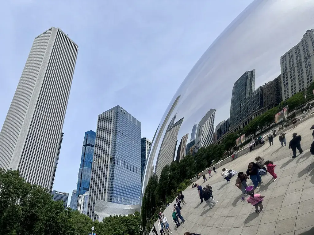 Eine spiegelnde Skulptur reflektiert hohe Gebäude der Skyline und eine Menschenmenge in einem Park. Der Himmel ist blau und leicht bewölkt, während die Architektur modern und variantenreich ist.
