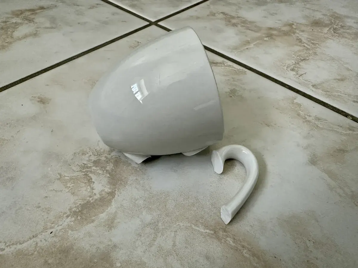 Eine gebrochene weiße Espresso-Tasse liegt auf einem gefliesten Boden. Der Henkel ist abgebrochen und die Tasse ist auf die Seite gefallen.