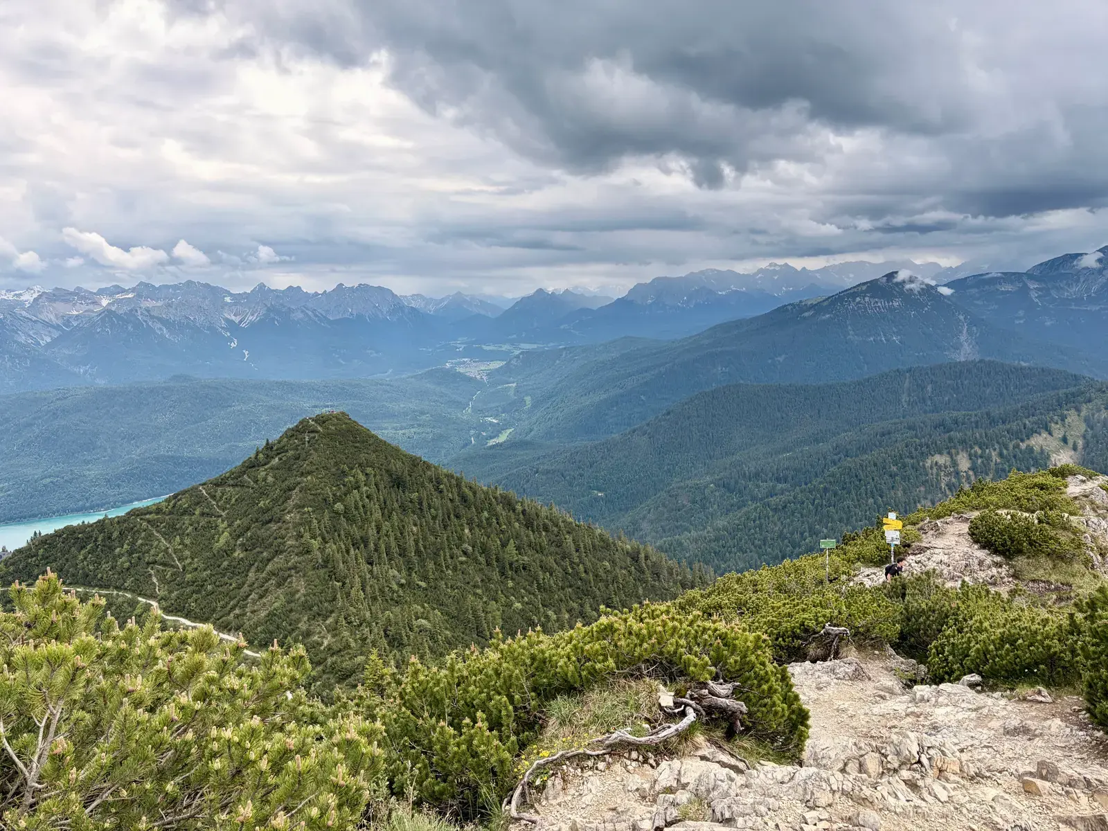 Blick über bewaldete Bergrücken und das Tal bis zu den Alpen. Dunkle Wolken ziehen über der Berglandschaft auf, links ist ein Stück des Kochlsees zu sehen.