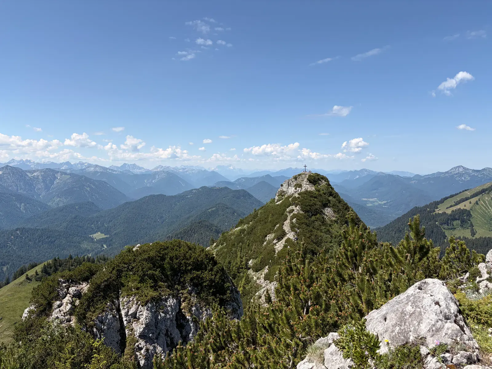 Blick vom Buchstein zum Roßkopf und über bewaldete Grate und Felsen in den Tegernseer Bergen. Im Hintergrund ziehen sich bei klarem Himmel mehrere Bergketten bis zum Horizont.
