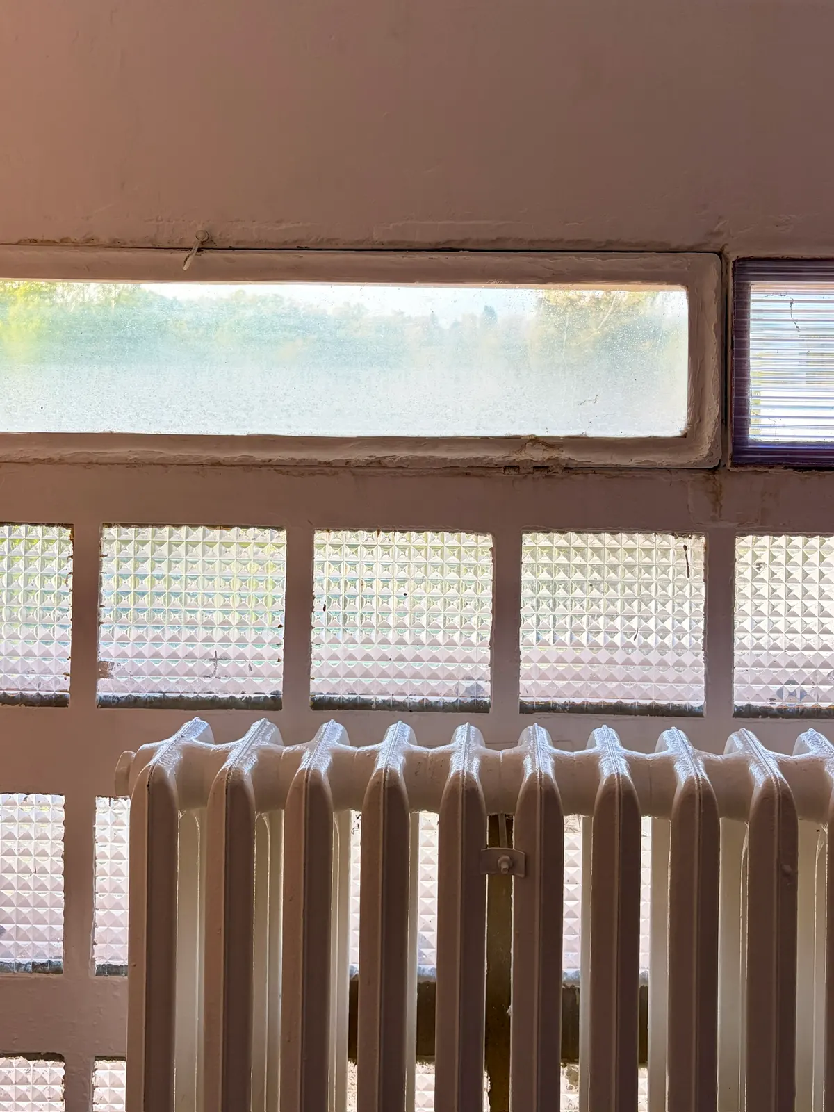 Alter Rippenheizkörper vor einer Wand aus Glasbausteinen und schmalen Oberlichtfenstern in einem Schulgebäude