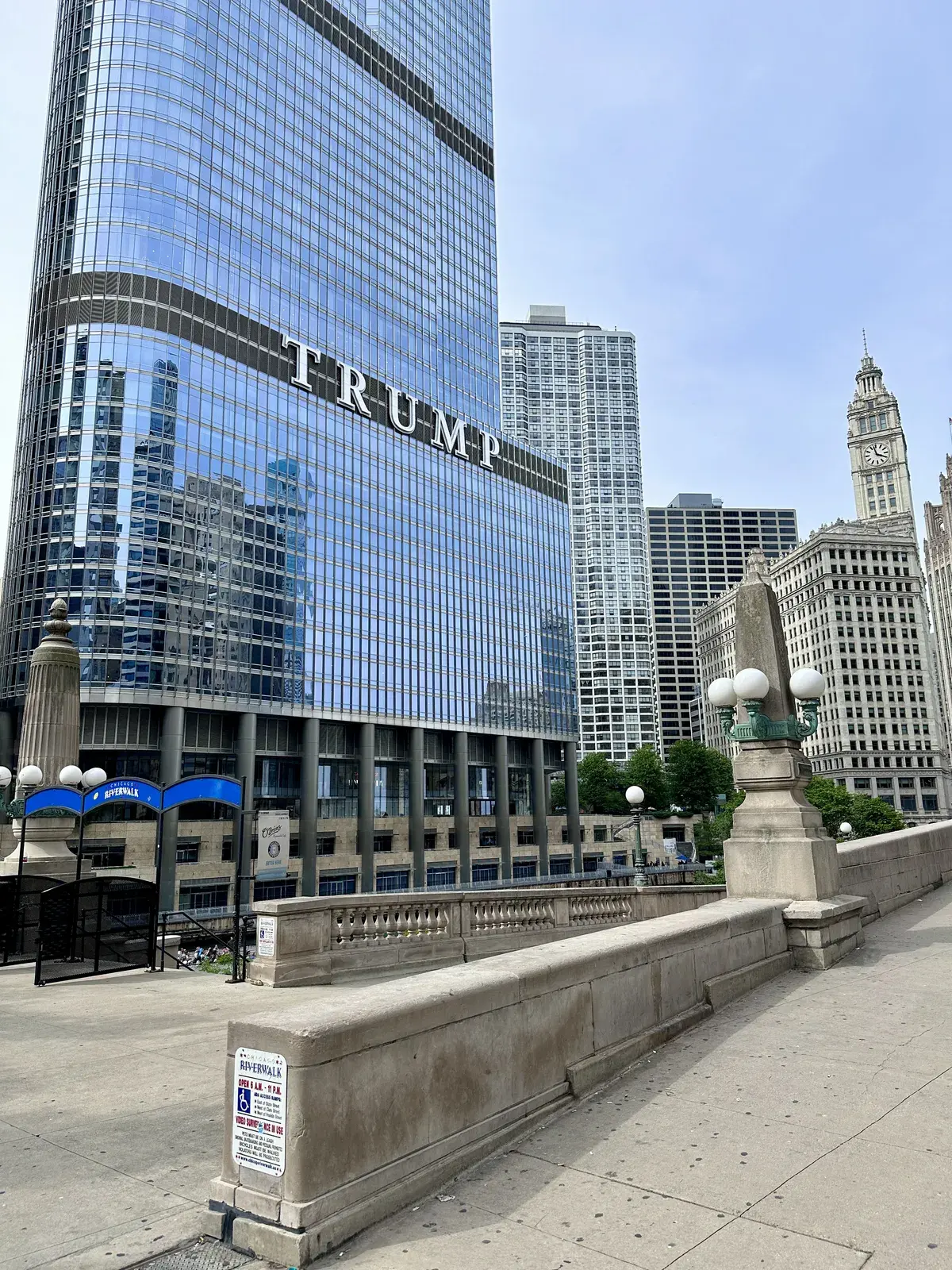 Das Bild zeigt das Trump International Hotel & Tower in Chicago, ein hochmodernes Wolkenkratzer-Gebäude mit einer reflektierenden Glasfassade. Im Hintergrund sind weitere moderne Wolkenkratzer und der historische Wrigley Building mit einem Uhrenturm sichtbar.