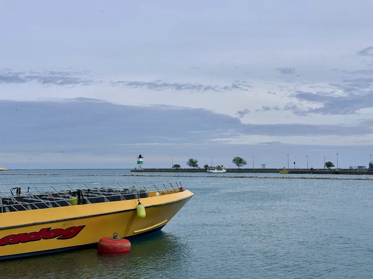 Ein gelbes Boot mit der Aufschrift „Speedog“ liegt im ruhigen Wasser, während im Hintergrund ein Leuchtturm und ein Hafen mit einigen Bäumen und einer Mauer sichtbar sind. Der Himmel ist bewölkt.