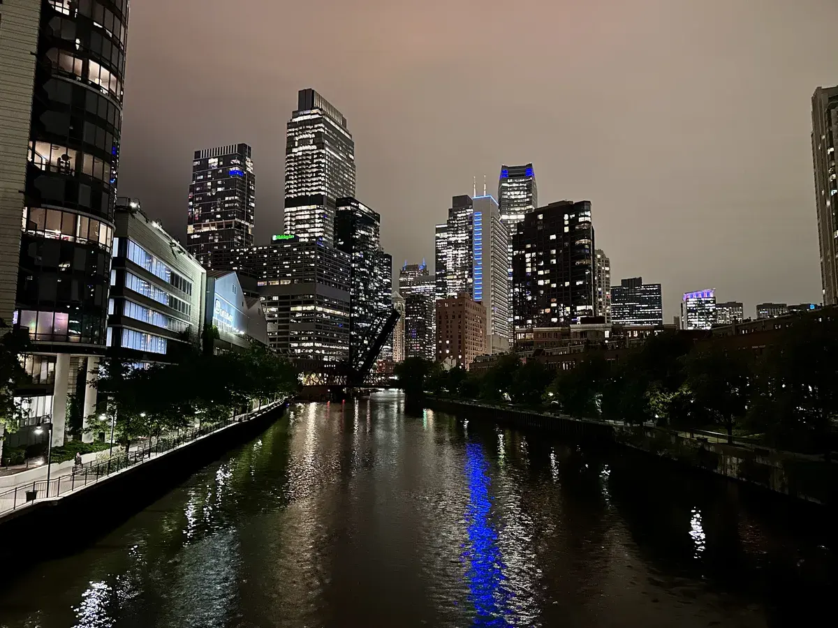 Nachtaufnahme eines urbanen Stadtbildes mit hohen Gebäuden, die in verschiedenen Farben beleuchtet sind. Im Vordergrund verläuft ein ruhiger Fluss, der das Licht der umliegenden Architektur reflektiert.
