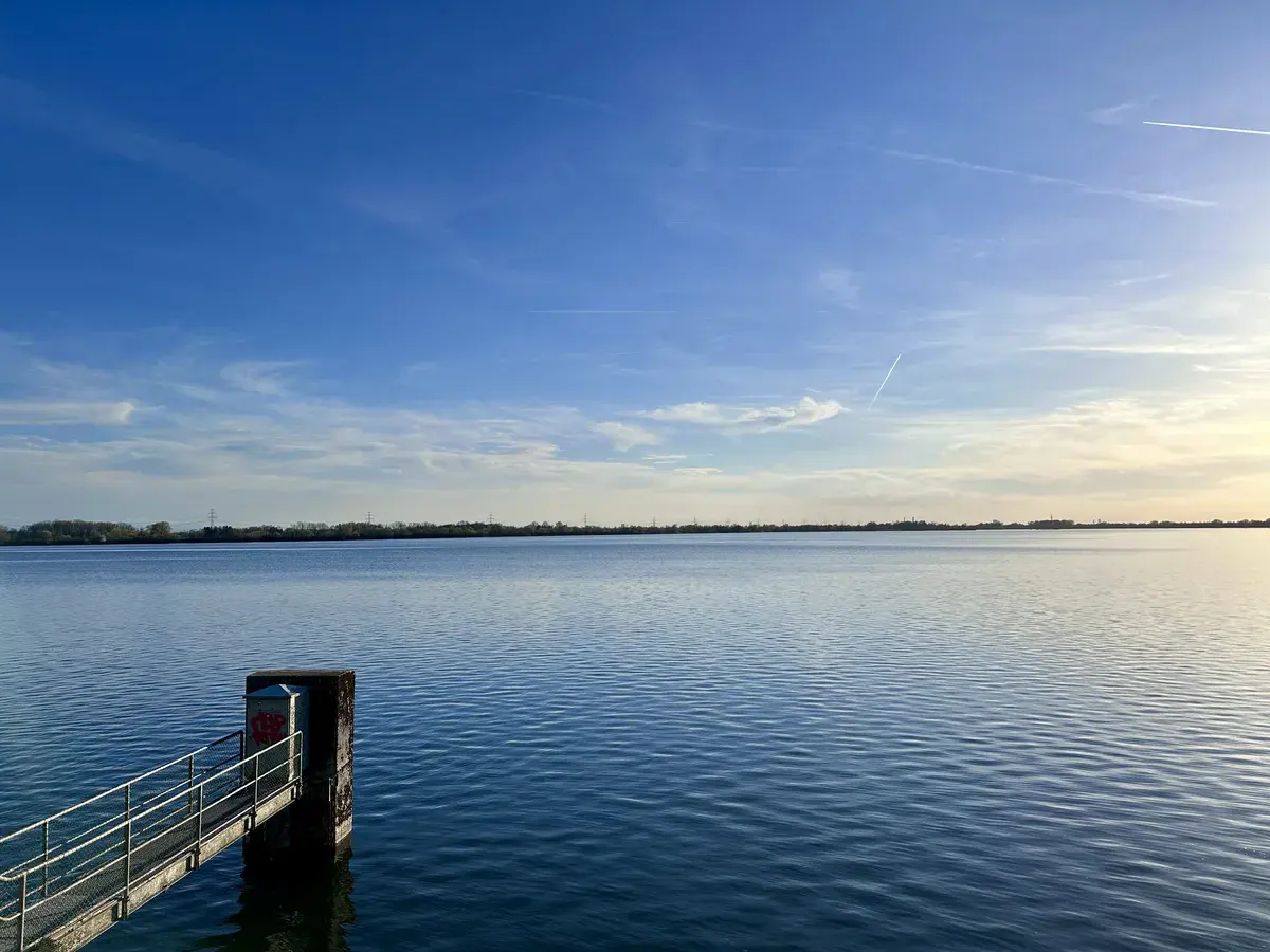 Ein ruhiger See unter einem klaren, blauen Himmel, mit sanften Wellen und einem kleinen Steg, der ins Wasser führt. Am Horizont sind Bäume und Strukturen zu erkennen.