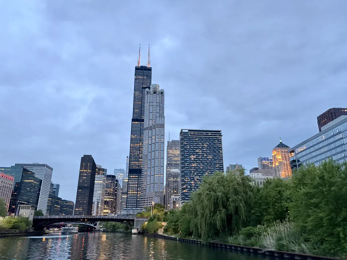 Die Skyline einer Stadt bei Dämmerung, mit hohen Wolkenkratzern, darunter der Willis Tower, der durch seine charakteristischen Spitzen hervorsticht. Im Vordergrund fließt ein ruhiger Fluss, umgeben von grünen Ufervegetationen.